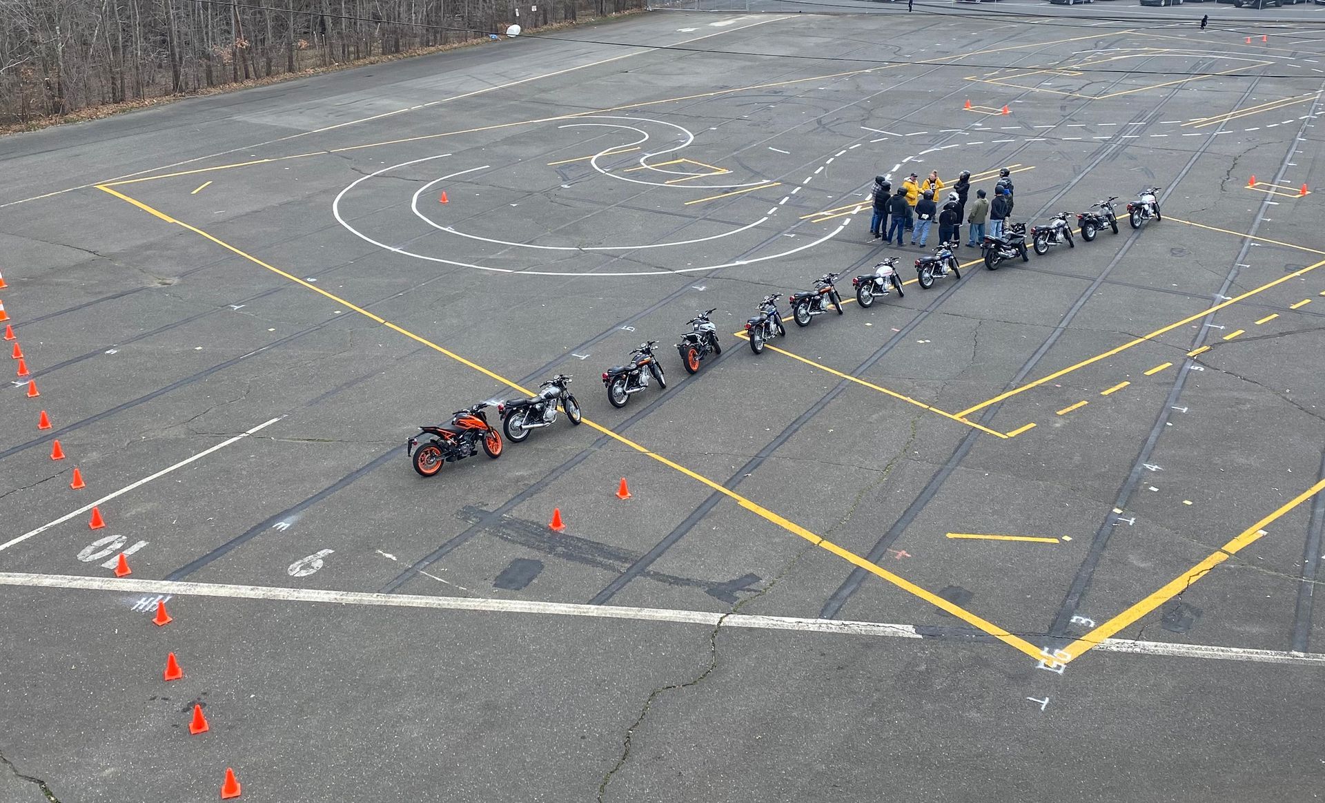 Motorcycle safety course: lined-up bikes, group of people, cones, and painted course markings on asphalt.