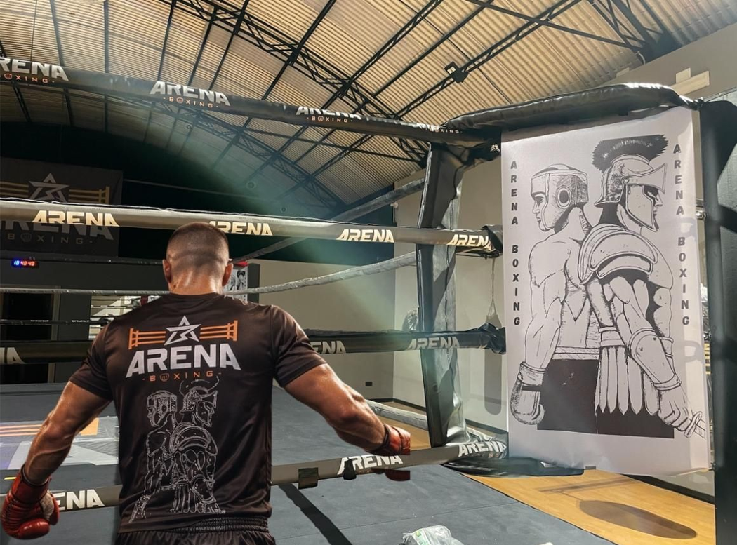 Un pugile su un ring sotto il tetto di uno stadio, di fronte a un grande bersaglio raffigurato su un poster appeso alle corde.