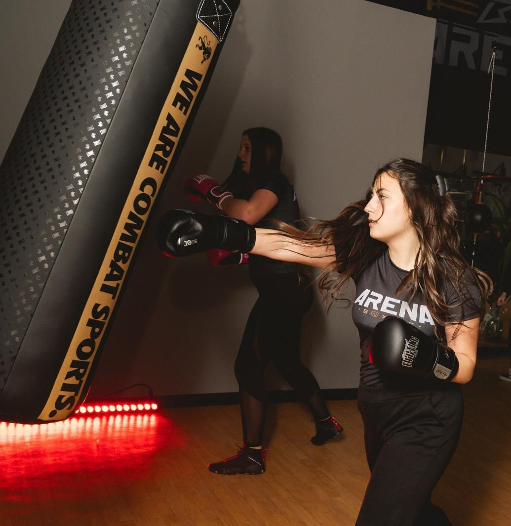 Una donna con guantoni da boxe neri colpisce un sacco da boxe in una palestra, illuminata da luci rosse sul pavimento.