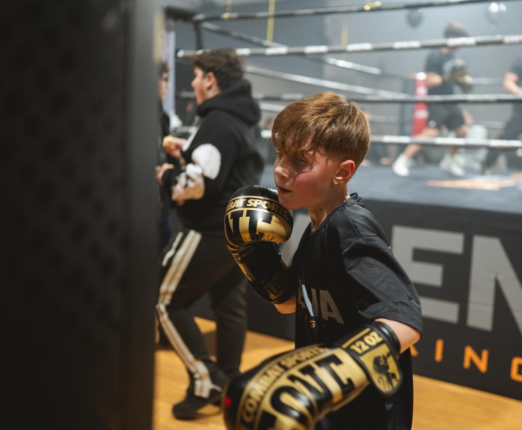 Giovane pugile con guantoni neri che si allena sul ring di una palestra, con un altro pugile sfocato sullo sfondo.