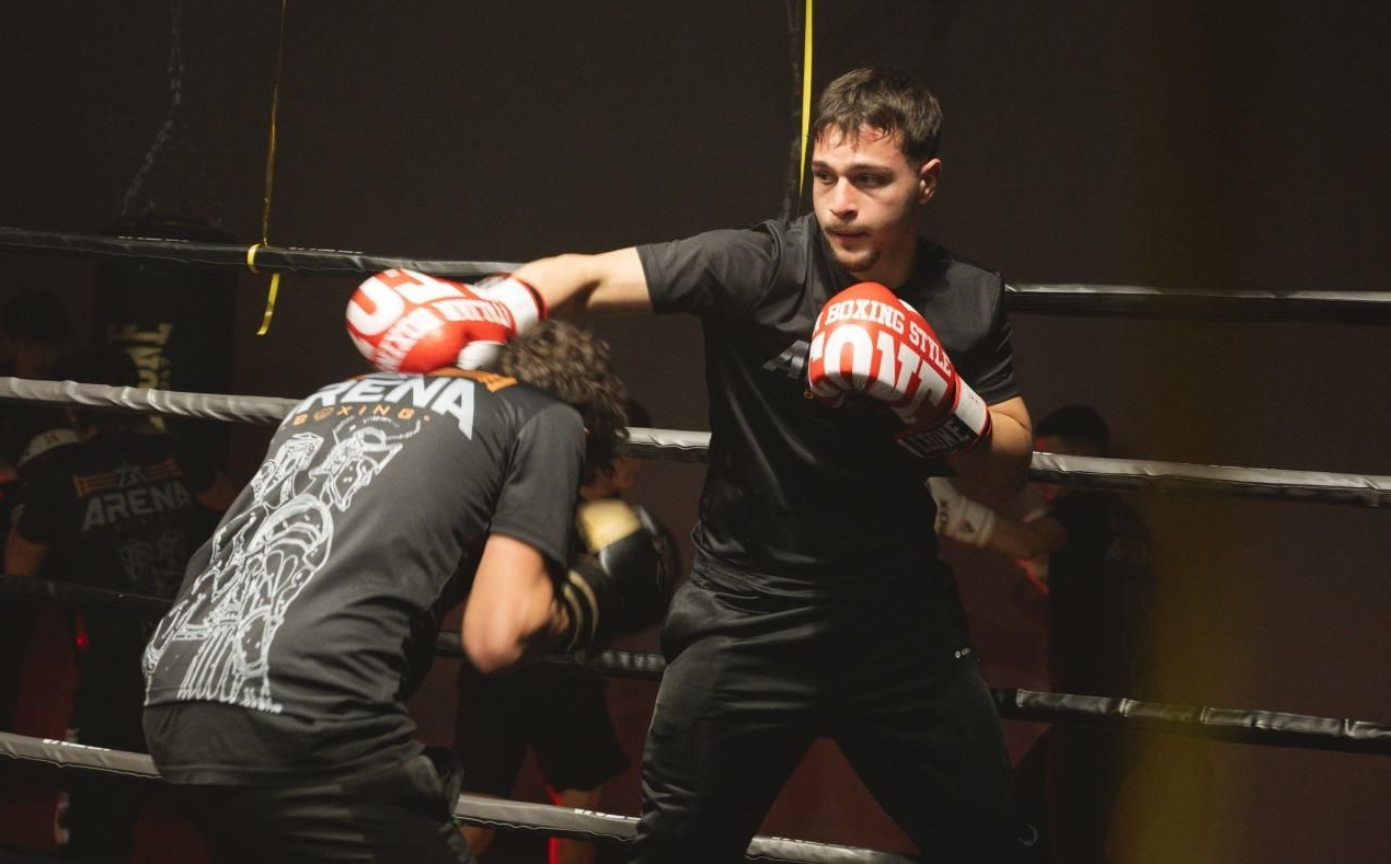 Due pugili si allenano su un ring, uno colpisce i guantoni mentre l'altro si prepara, in una palestra poco illuminata.