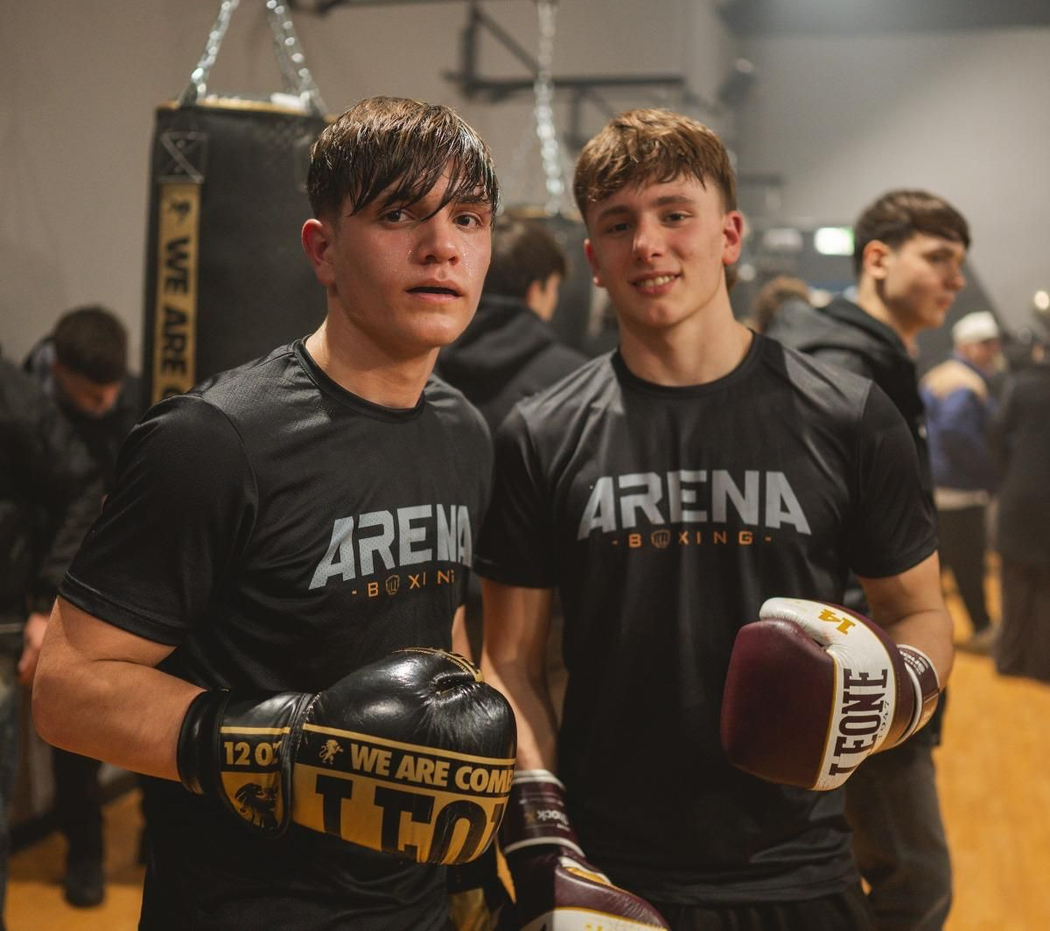 Due allievi di boxe, con indosso magliette e guantoni neri della ARENA, posano sul ring di una palestra.