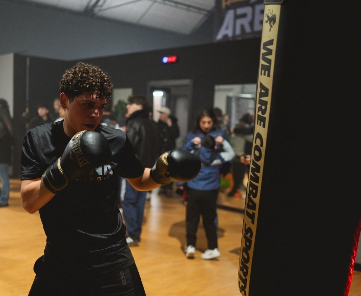 Un pugile con i guantoni neri si allena in palestra, con un sacco da boxe e degli spettatori sullo sfondo.