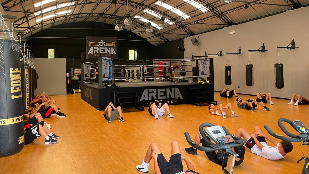 Persone che si allenano su dei tappetini in una palestra al coperto con un ring da boxe e pareti nere e arancioni.