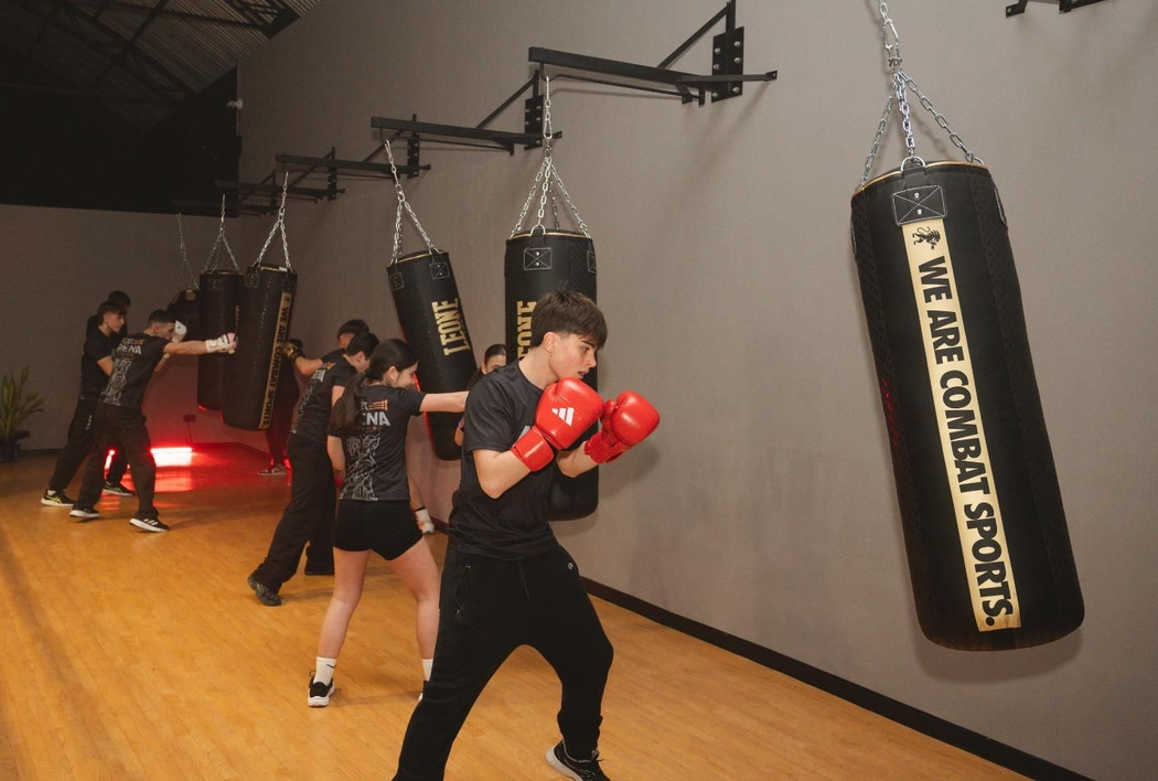 Persone che si allenano con la boxe in una palestra, con sacchi da boxe appesi e guantoni rossi su un pavimento di legno.