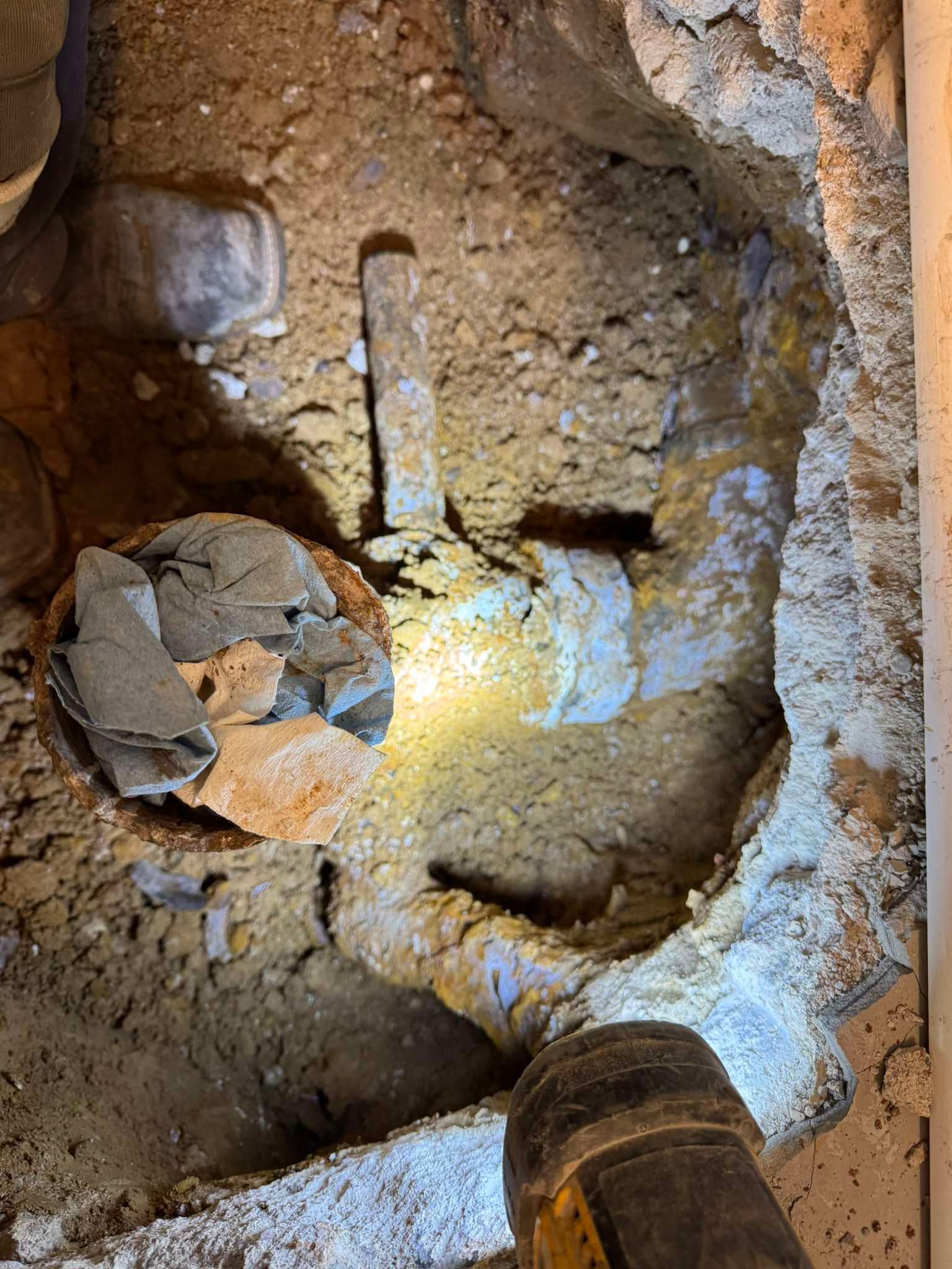 A hole dug into dirt floor revealing exposed, damaged plumbing pipes lit by a flashlight.