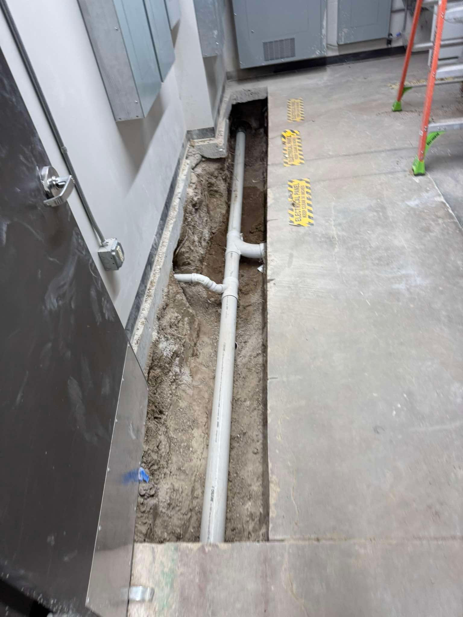 A trench cut into a concrete floor reveals a white PVC pipe assembly inside an industrial building.