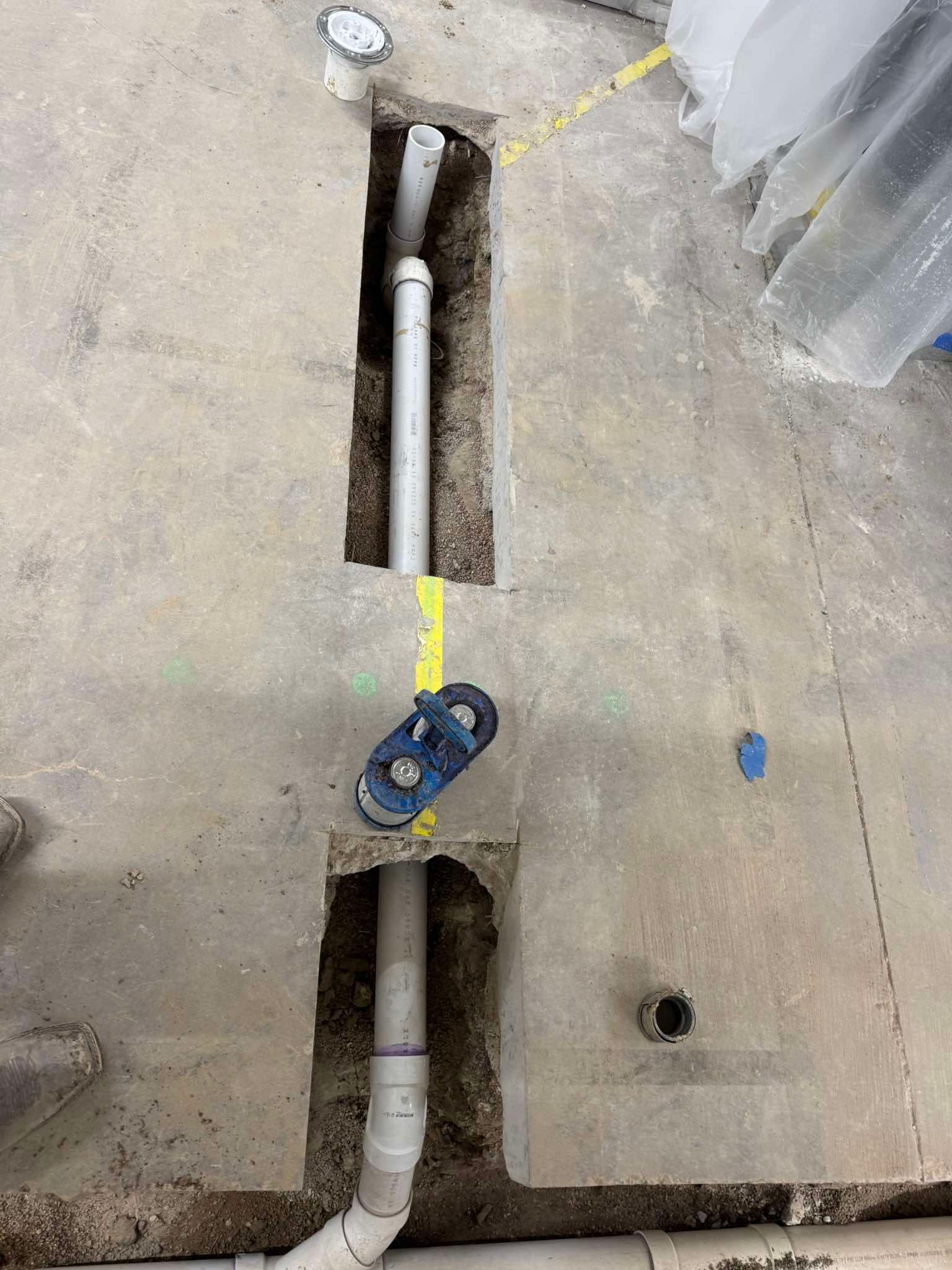 Two channels cut into a concrete floor reveal PVC plumbing pipes with a blue tool resting on the surface between them.
