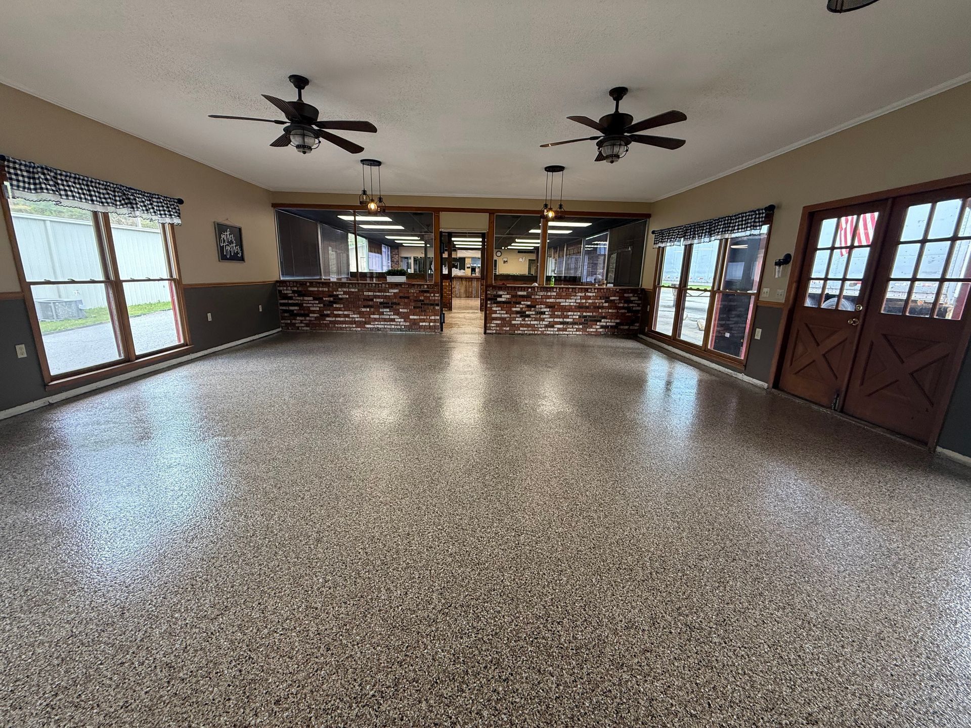 Spacious room with speckled epoxy flooring, two ceiling fans, large windows, and a dark wood door.