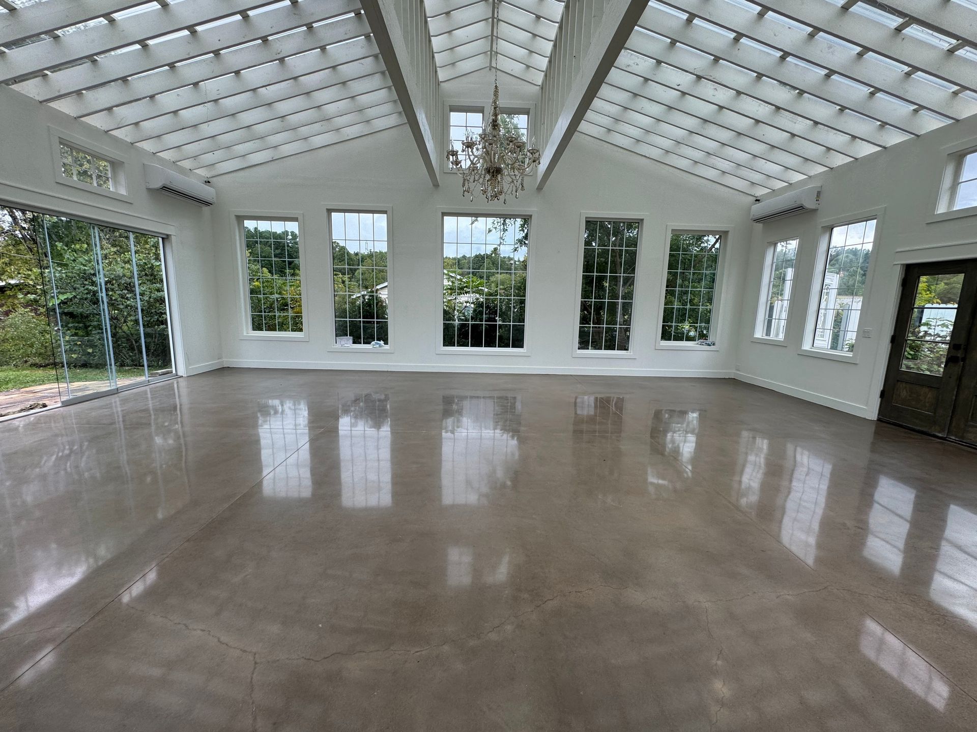 A sunlit, empty event space with a vaulted glass-panel ceiling, white walls, large windows, and polished concrete floors.