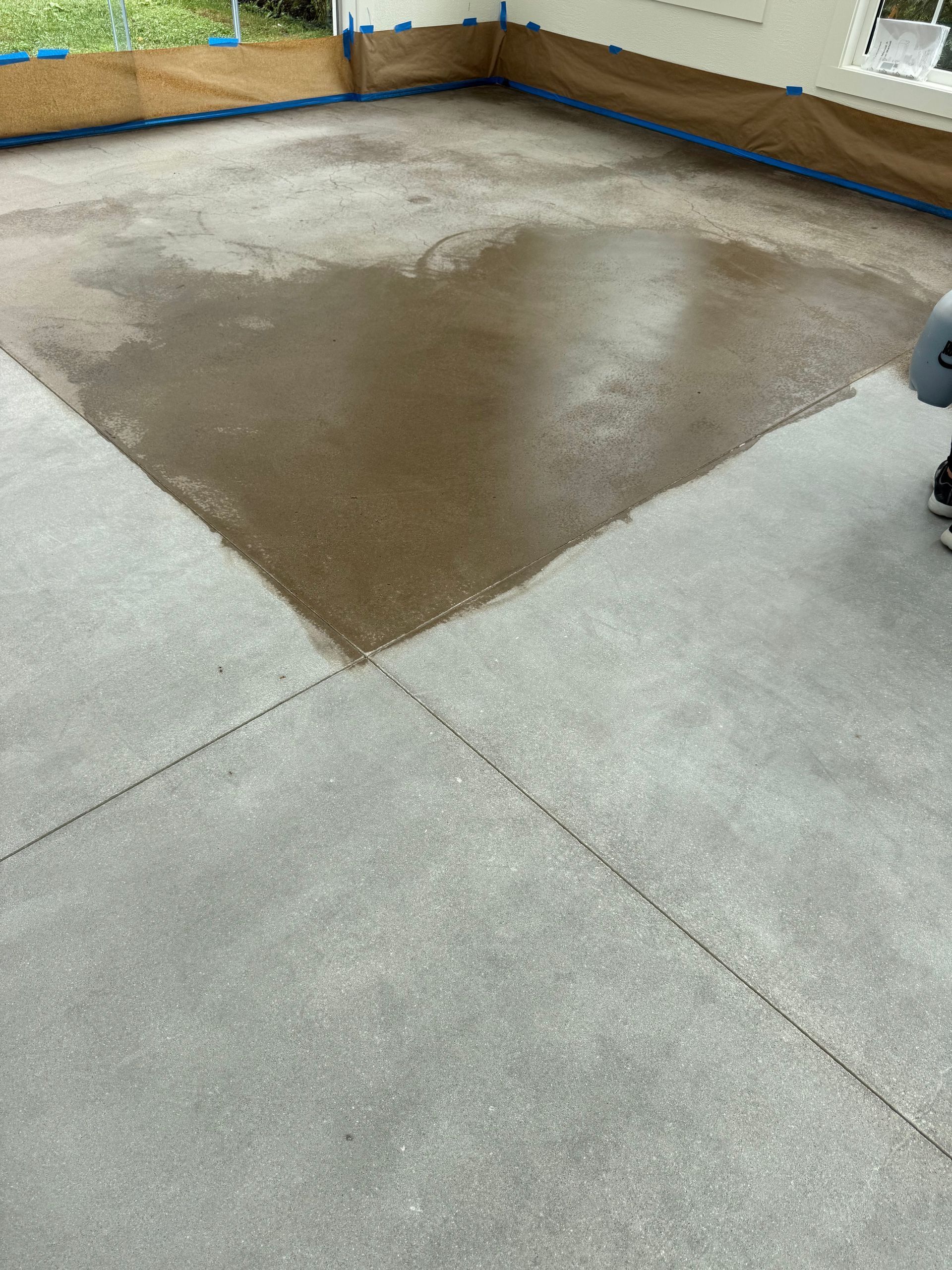 Concrete floor with a large, dark, damp patch in the center, surrounded by taped-off walls in a room under renovation.