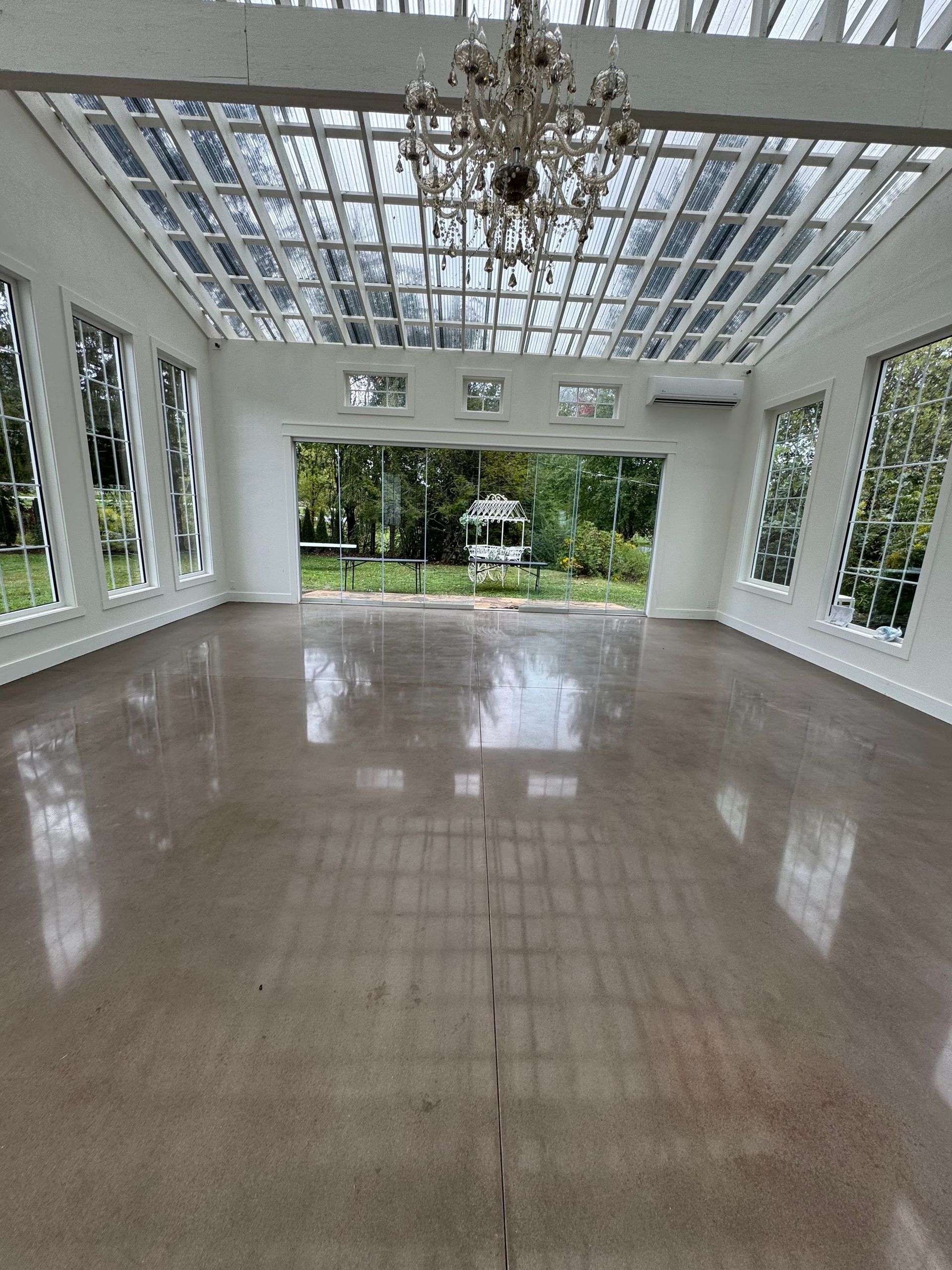 An empty, bright event hall with glass walls, a high glass ceiling, a central chandelier, and a polished concrete floor.