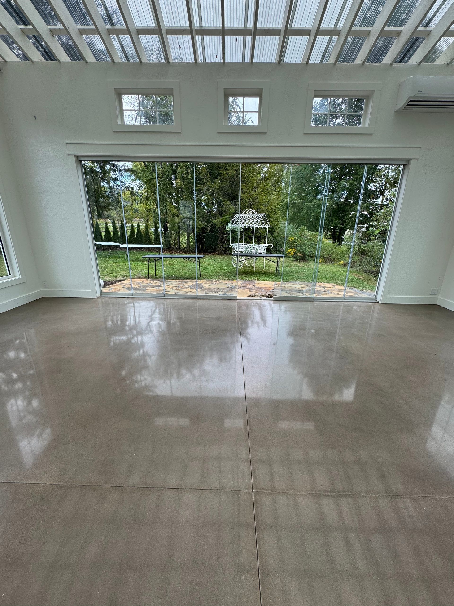 A wide, polished concrete floor inside a bright room, looking out through a large open wall to a grassy garden.