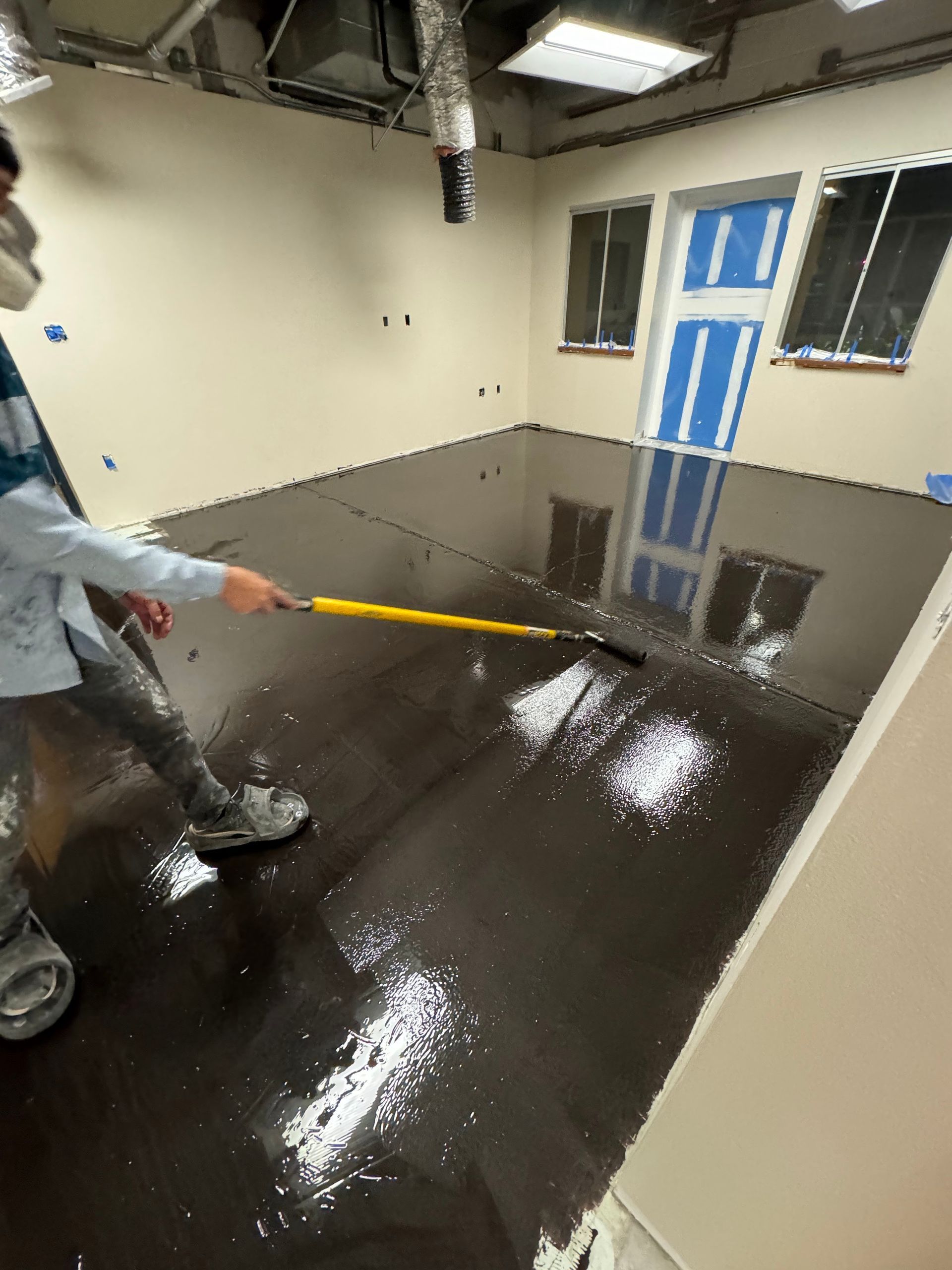 A worker uses a squeegee to spread glossy dark brown epoxy coating across a room's floor.