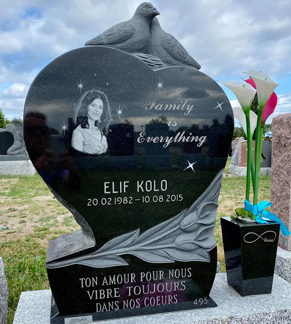 Pierre tombale noire en forme de cœur avec une photo, des colombes et une inscription en français ; à côté, un pot de fleurs.