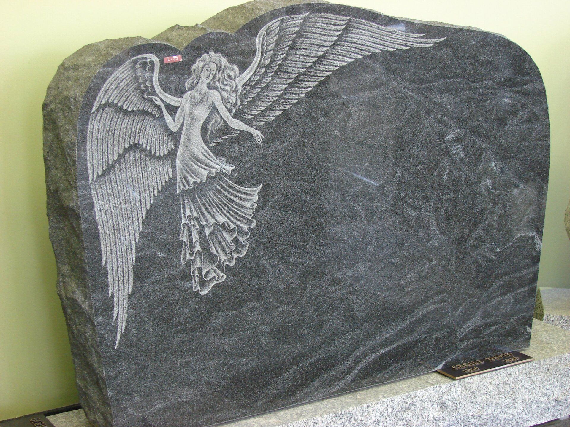 Une pierre tombale en granit gris foncé ornée d'un ange gravé aux grandes ailes finement détaillées qui s'étendent sur toute la surface de la pierre.
