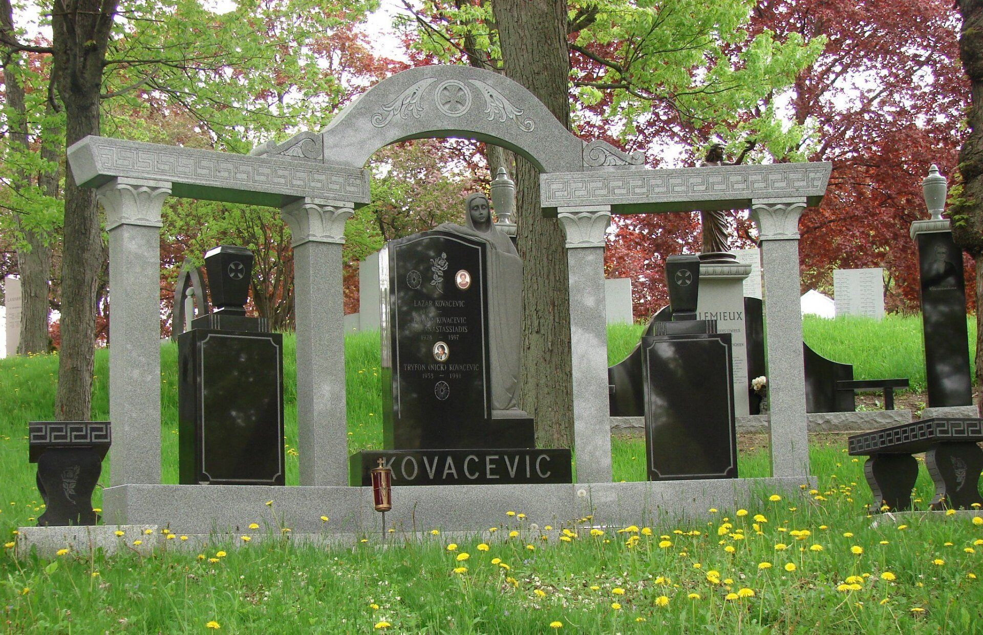 Un monument élaboré en pierre grise, en forme d'arche, avec trois pierres tombales en granit noir et un banc, dans un cimetière herbeux.