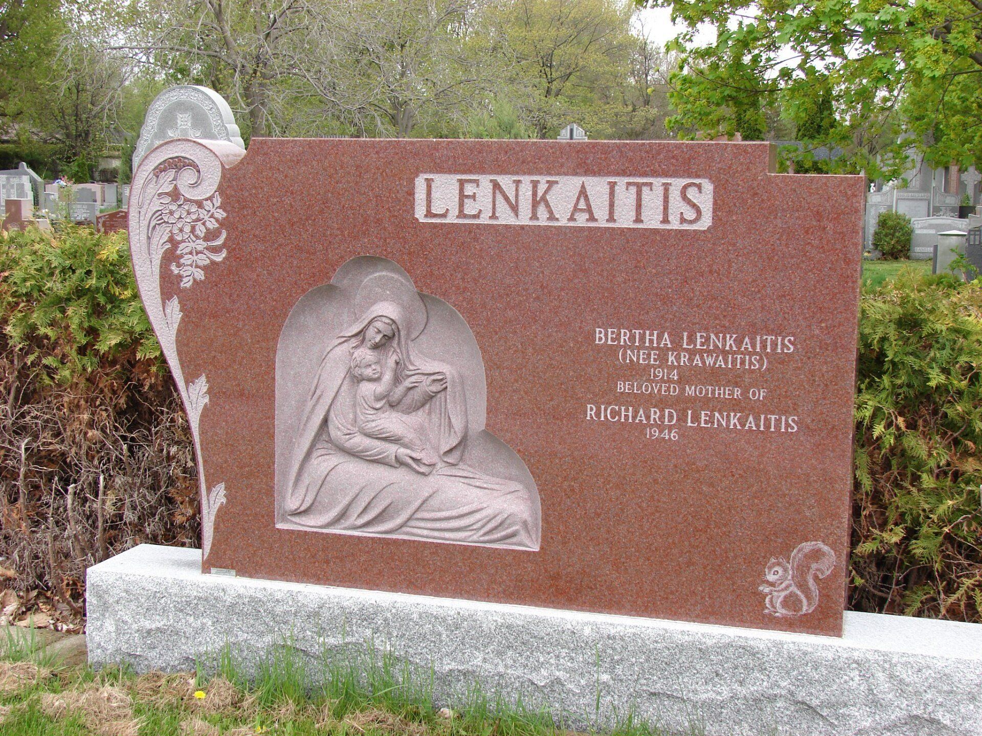 Pierre tombale en granit rouge ornée d'une image sculptée de la Vierge Marie et de l'Enfant, portant l'inscription « LENKAITIS », dans un cimetière.