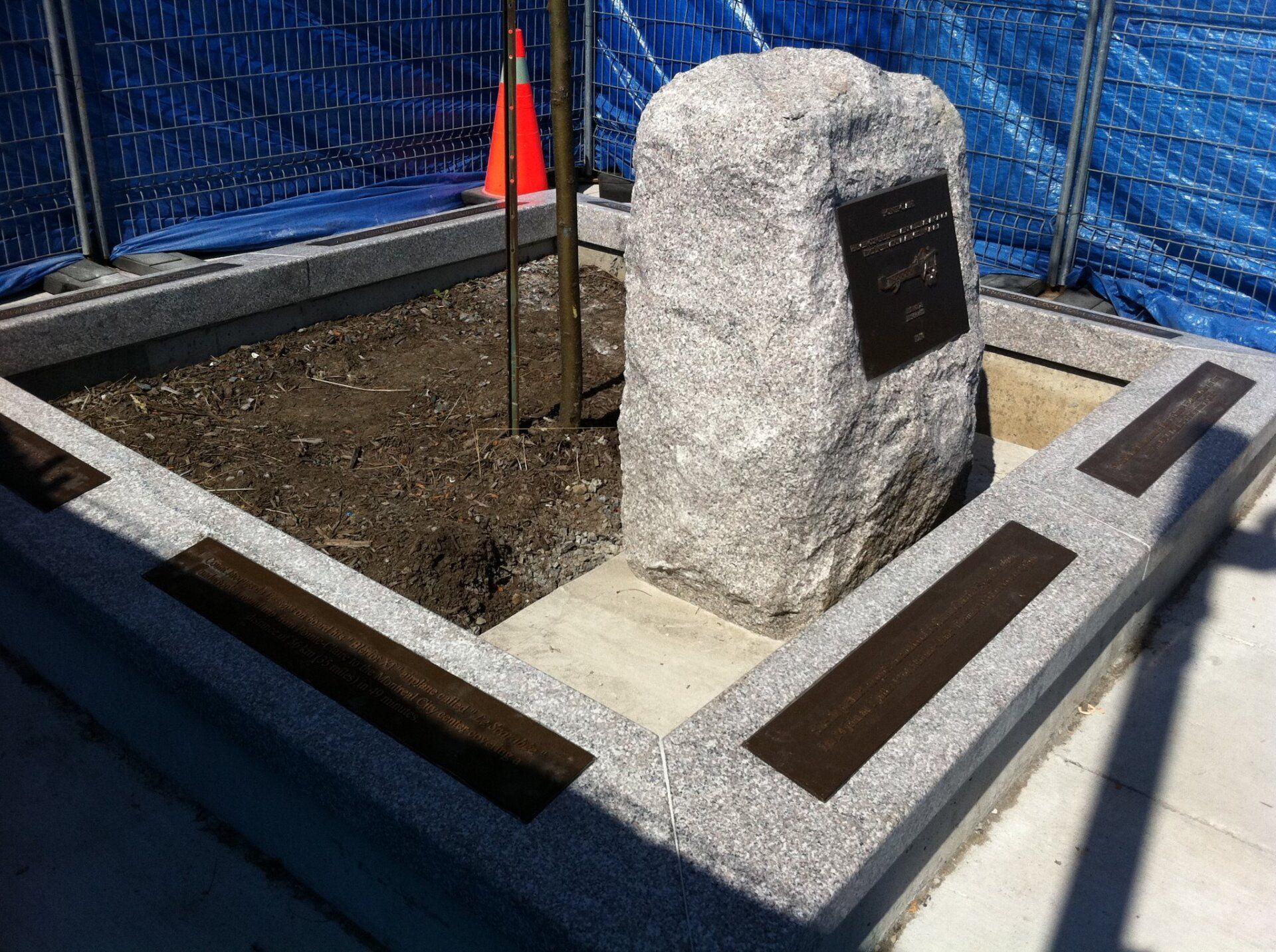 Stone monument with plaque in a planter box, small tree, dark metal plaques along edge.