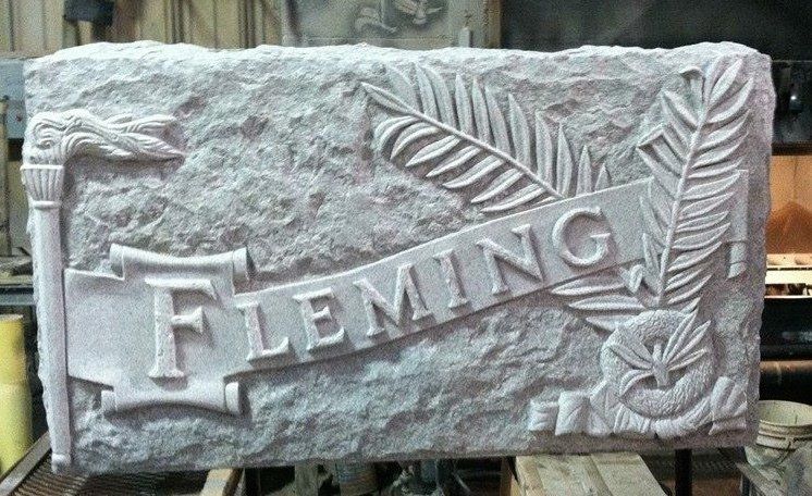 Sculpture en pierre grise portant l'inscription « FLEMING » en lettres en relief sur une bannière ornée de plumes et d'une torche.