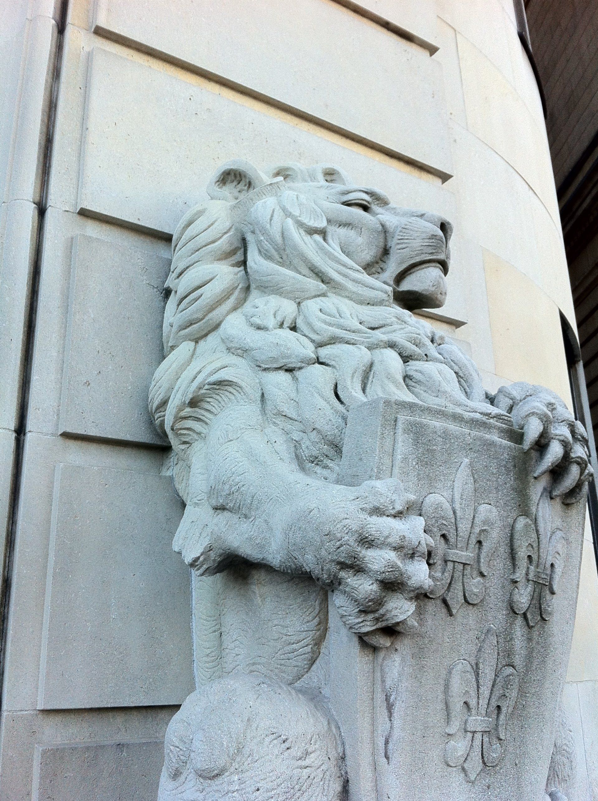 Stone lion statue holding a shield. It is on a building corner.