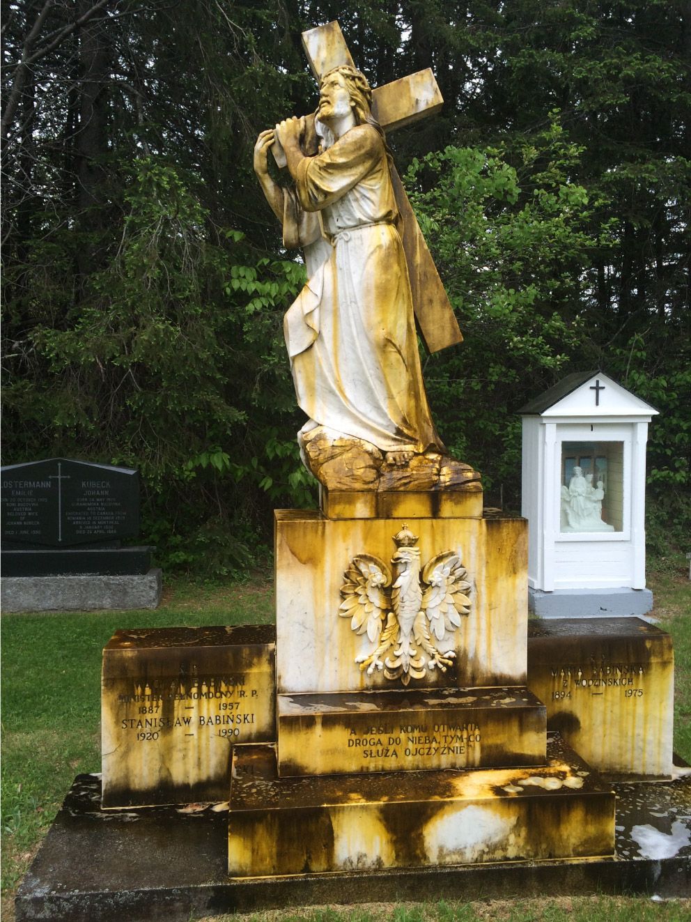 Statue of Jesus carrying a cross; in a cemetery. Yellowing stone, white robes, cloudy sky, small chapel nearby.