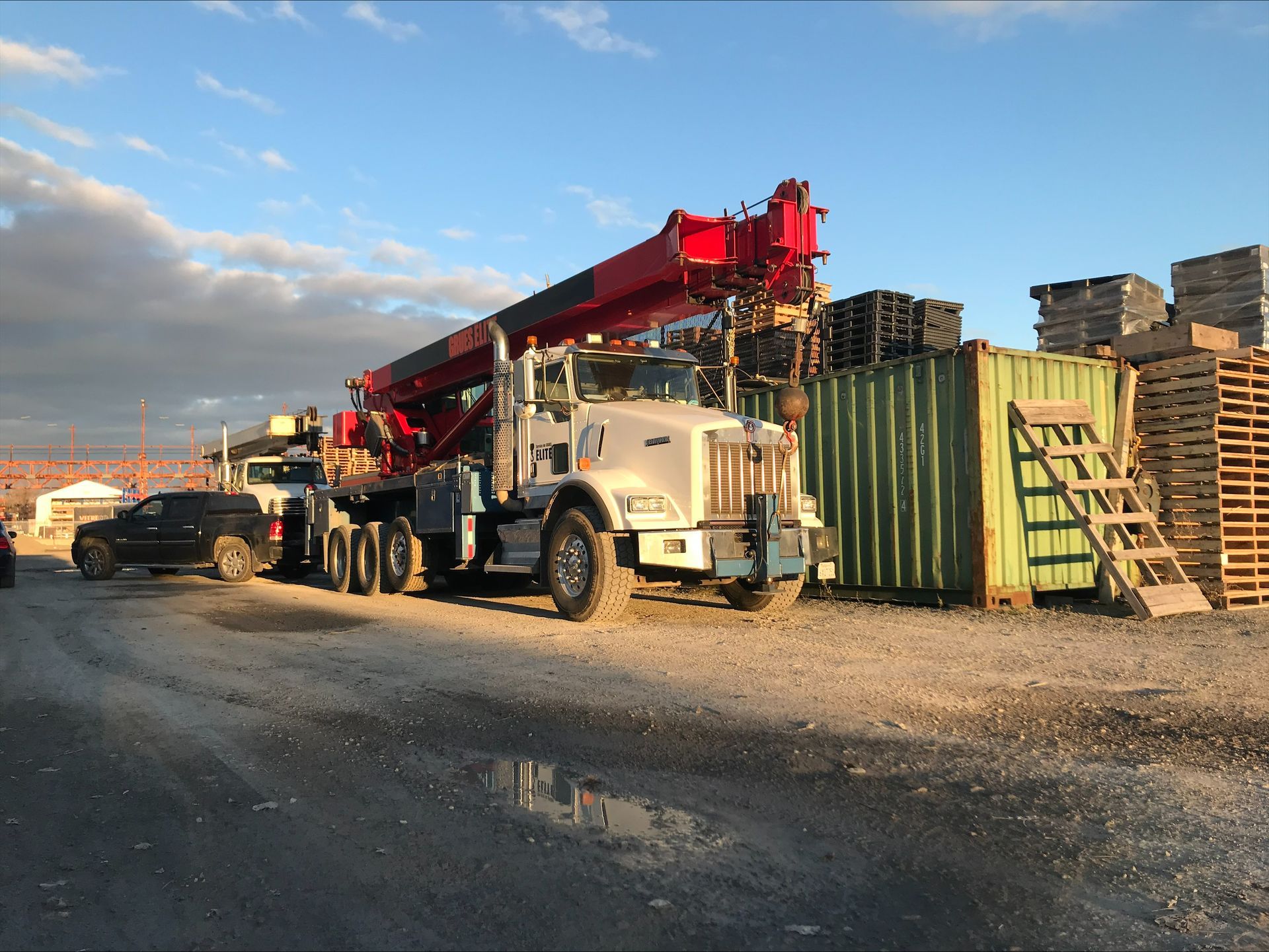 Un gros camion-grue avec sa flèche déployée se tient près de conteneurs et de palettes sur un terrain en gravier, sous un ciel nuageux.