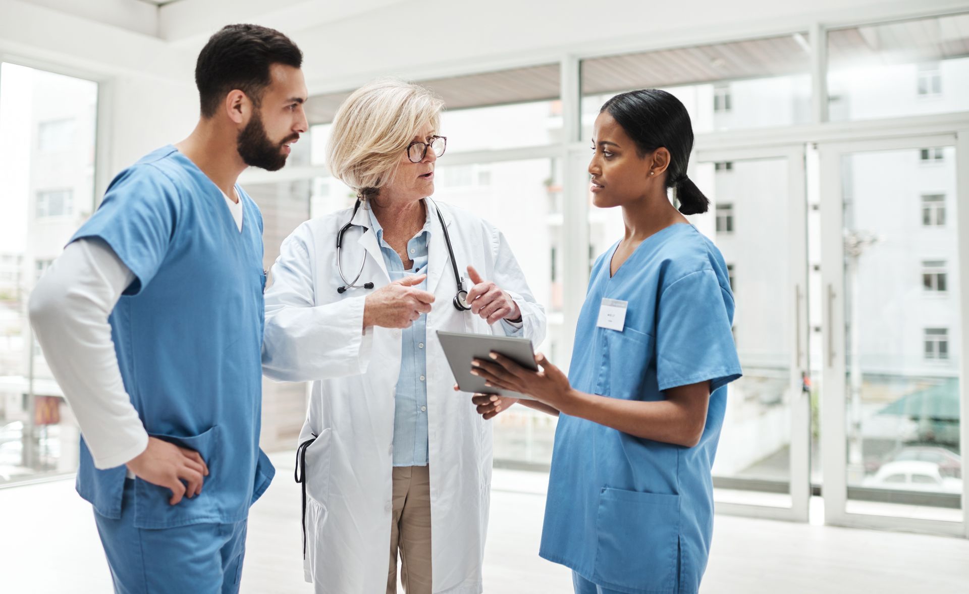 Maria Lewis Two medical staff in blue scrubs and a physician in a white coat consult while looking at a tablet in a bright hospital.