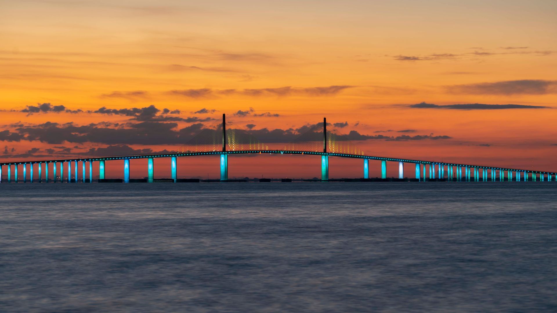 Wide lit bridge at sunset over calm water, with orange sky and clouds.