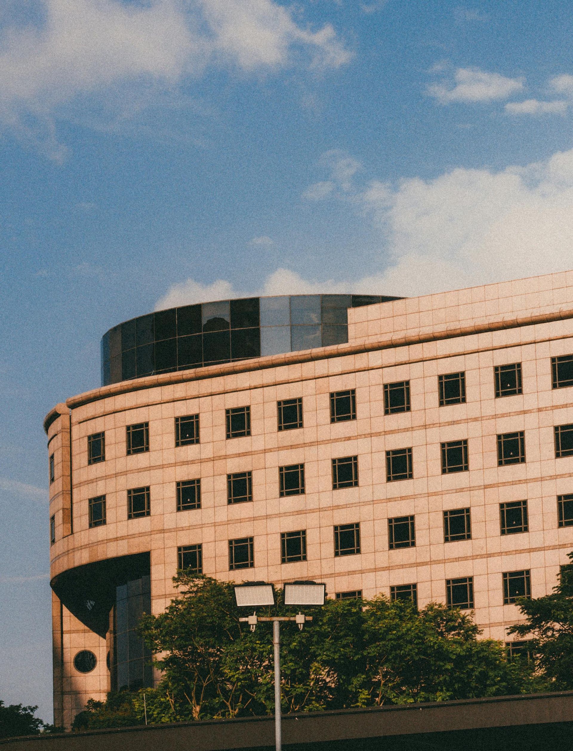 Curved beige building with small square windows, topped by a dark circular structure, against a blue sky.