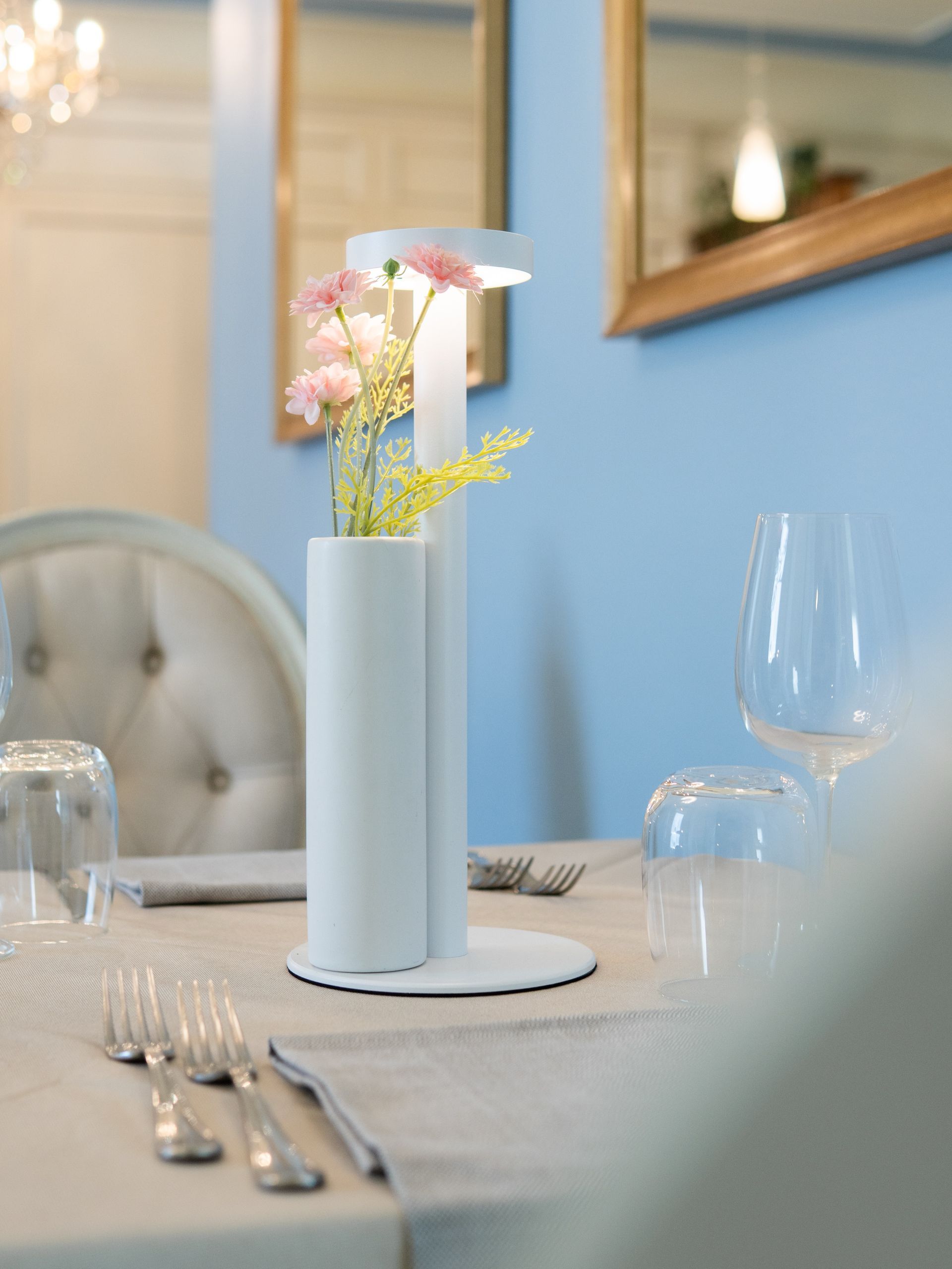 Un vaso bianco con fiori rosa su un tavolo da pranzo apparecchiato in una stanza illuminata da una luce soffusa.