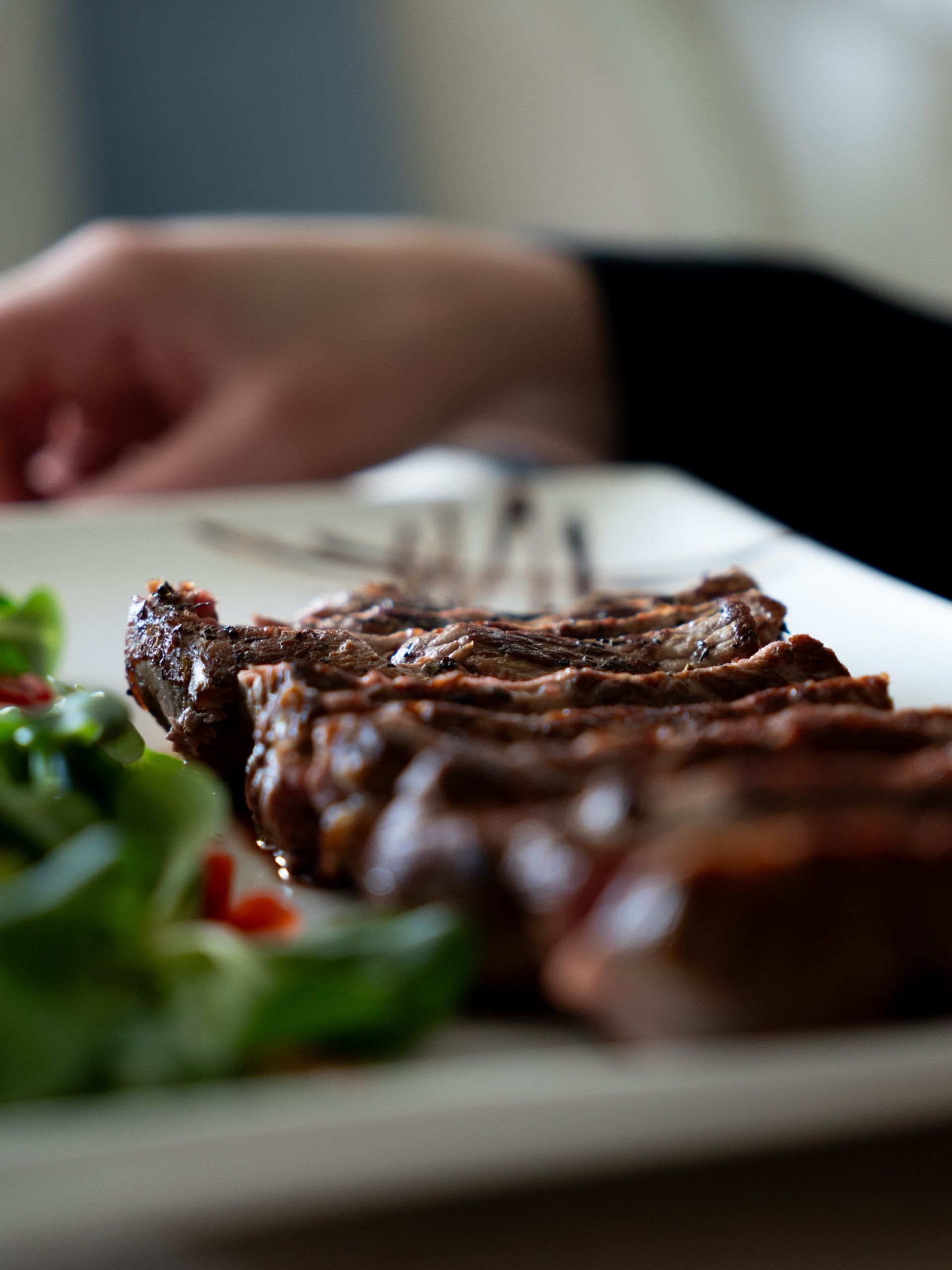 Bistecca alla griglia su un piatto bianco con insalata di contorno, con una mano sfocata sullo sfondo.