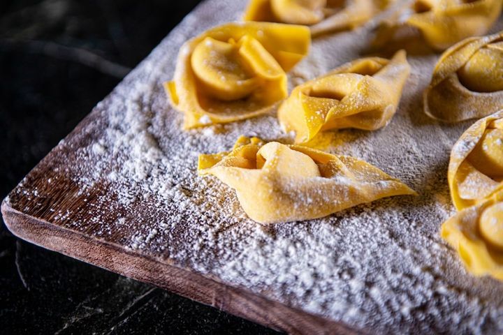Tagliere di legno infarinato con tortellini crudi, illuminato da una luce calda su uno sfondo scuro.