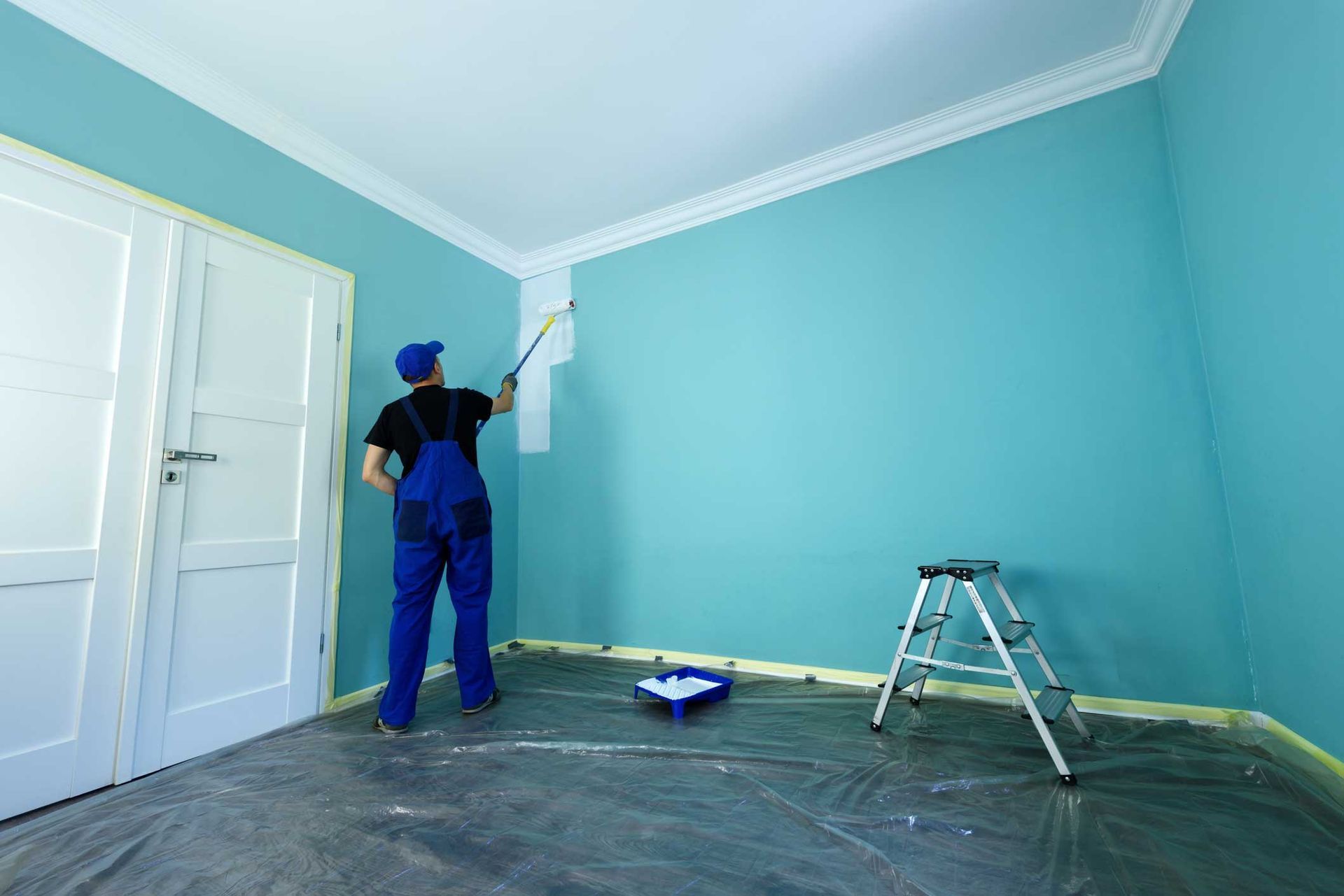 Person in blue overalls painting a light blue wall white near the ceiling trim, with a roller and ladder in the room.