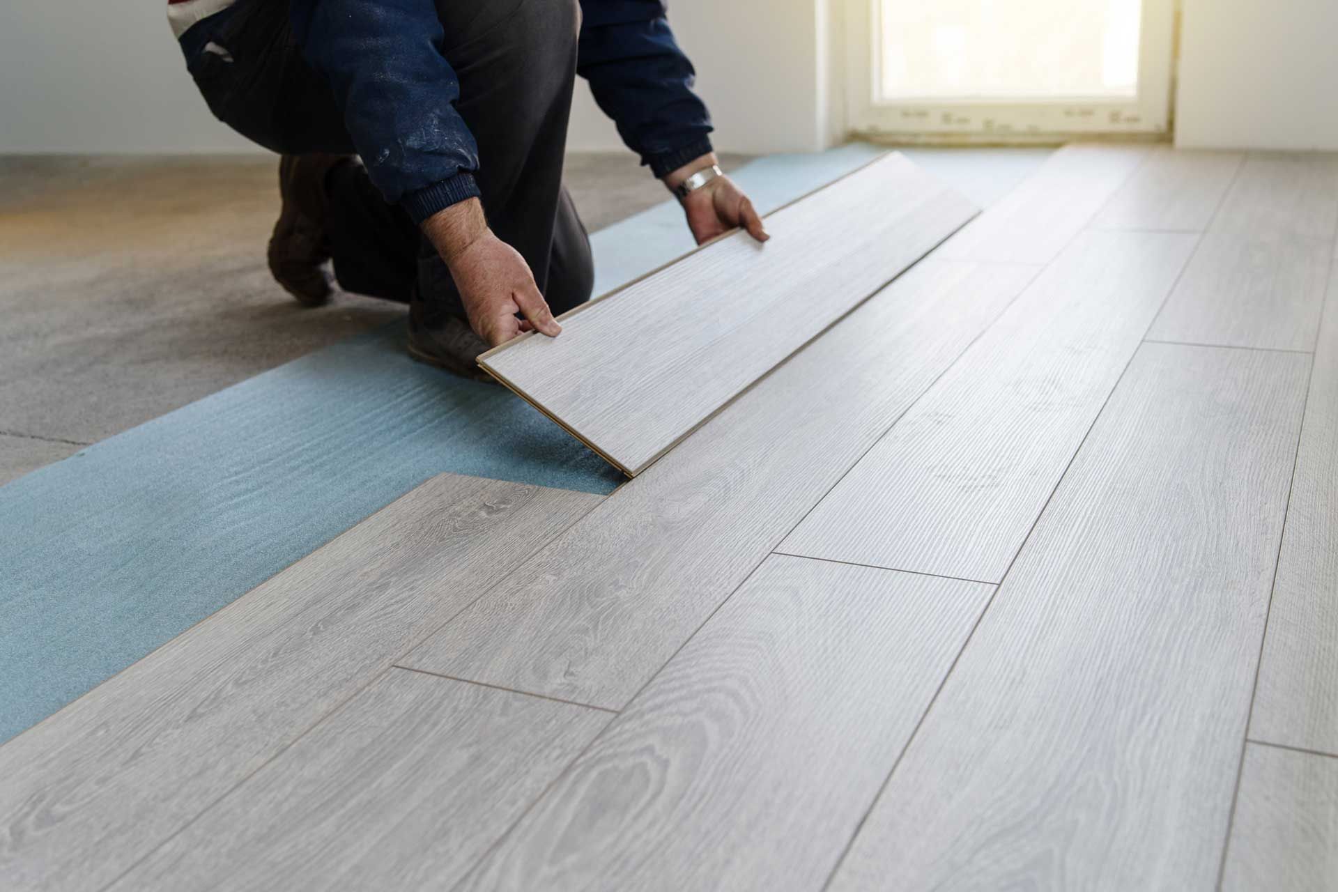 Person kneeling, installing light gray wood flooring. Flooring planks are being placed on blue underlayment.