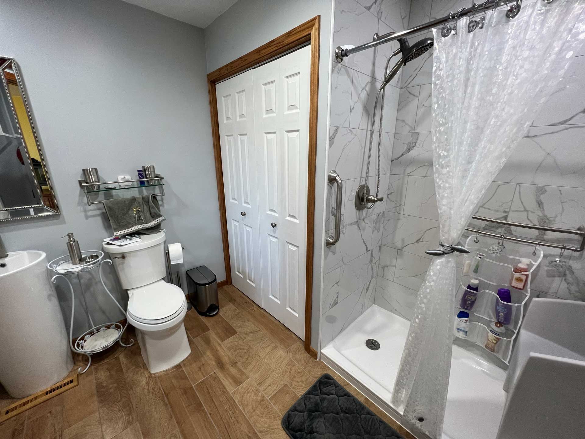 Bathroom with a toilet, pedestal sink, shower with a grab bar, and closed bi-fold door. Bathroom with a toilet, pedestal sink, shower with a grab bar, and closed bi-fold door.