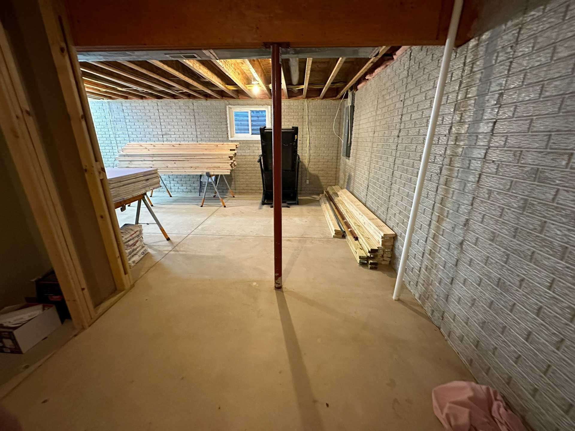 Basement with concrete floor, brick-patterned walls, wooden beams, a support post, and construction materials. Basement with concrete floor, brick-patterned walls, wooden beams, a support post, and construction materials.