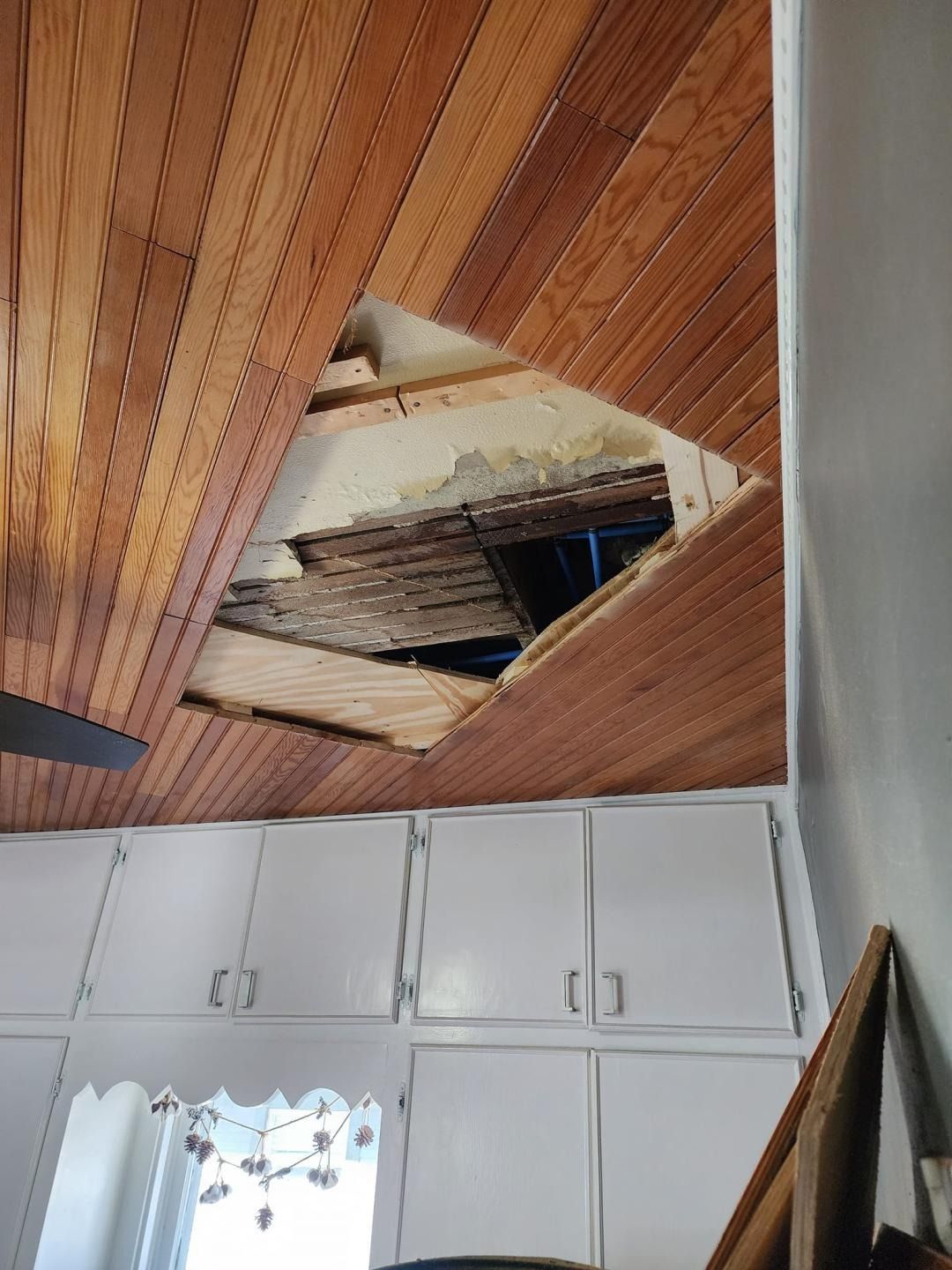 Damaged wooden ceiling with a large hole above white cabinets in a room