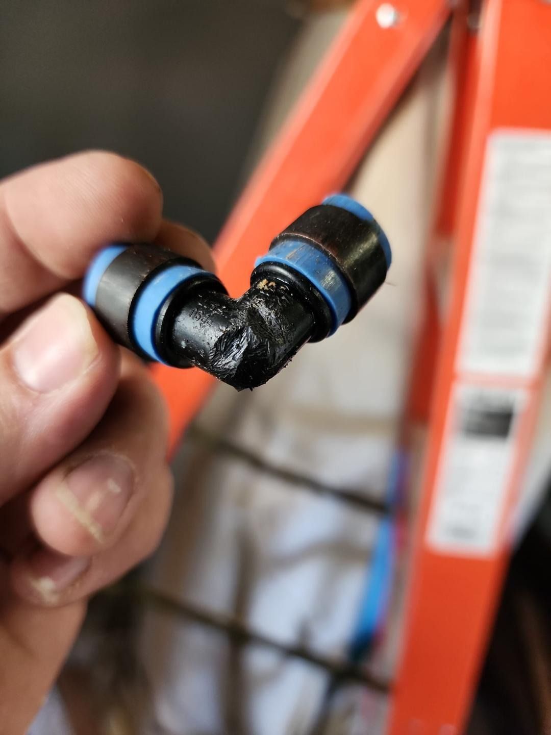 Hand holding a black and blue elbow connector with a corroded joint in front of an orange ladder