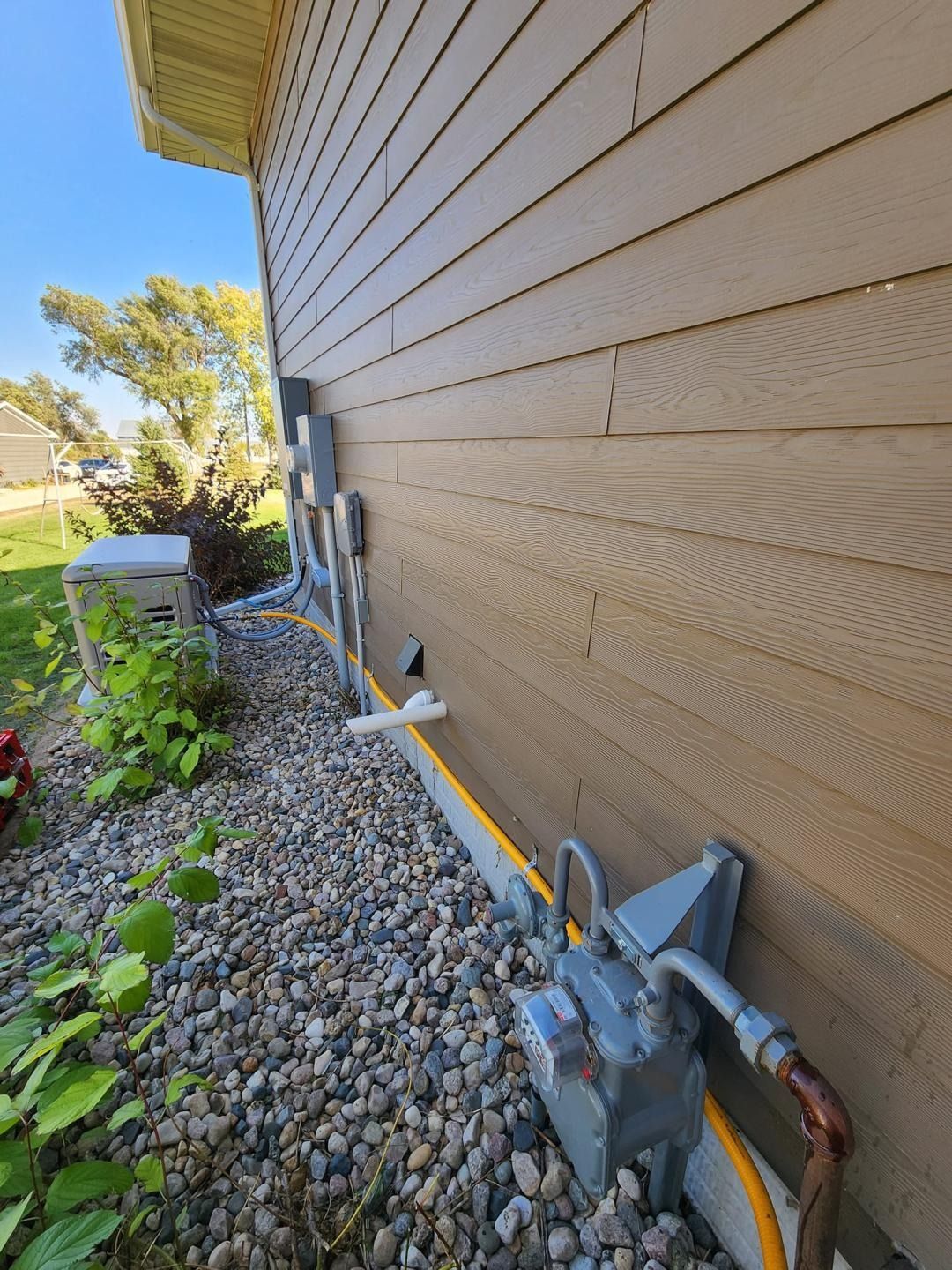 Side view of a house with utility meters, pipes, gravel, and a yellow cable along the foundation.
