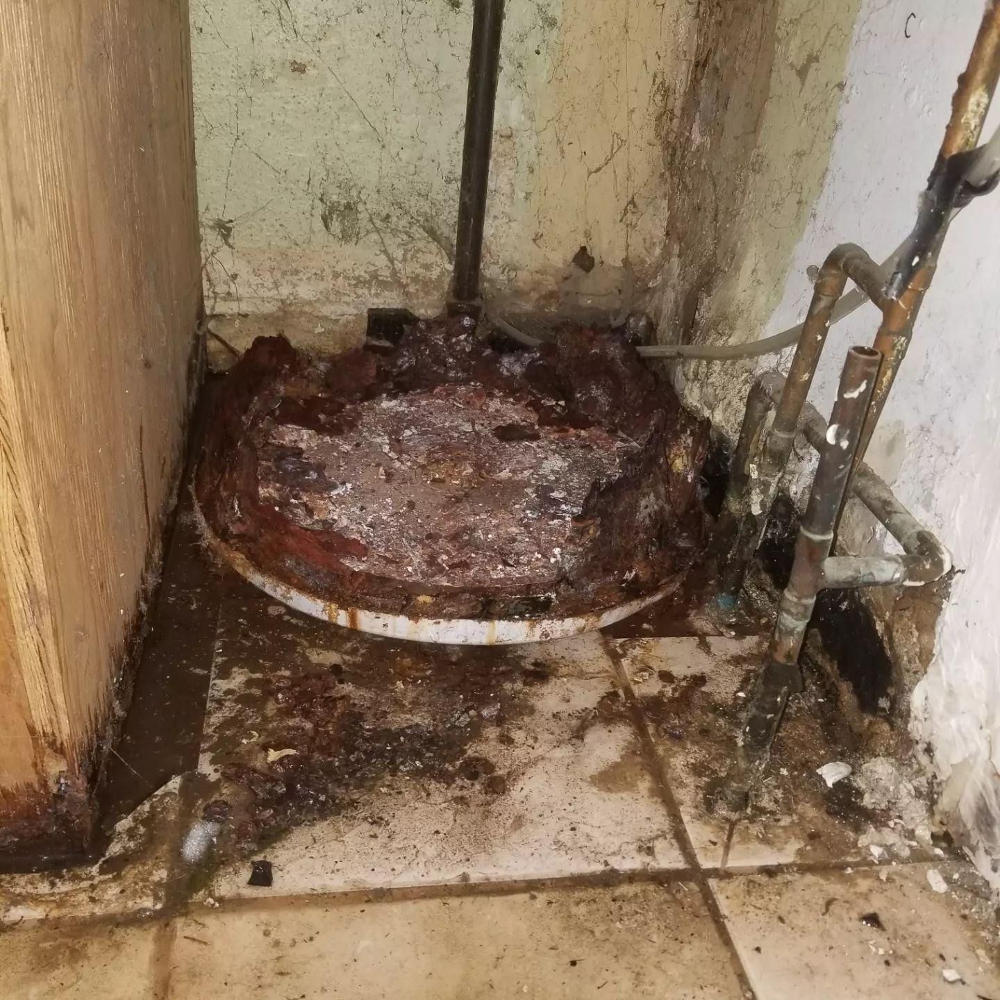 Rusty, heavily corroded metal tank in a dirty, stained utility corner with exposed pipes.