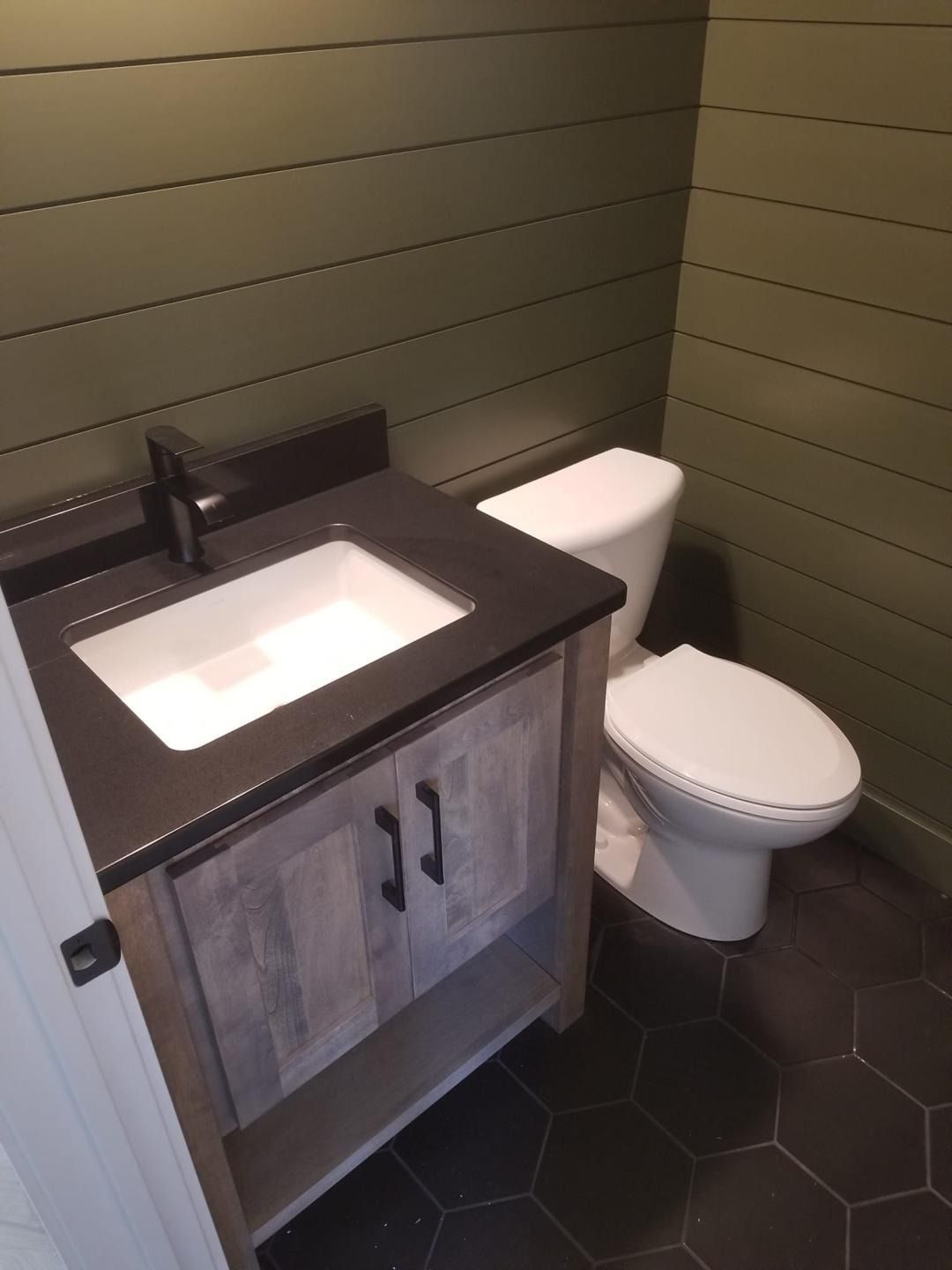 Small bathroom vanity with sink beside a toilet, dark countertop, wood cabinet, and hex tile floor.