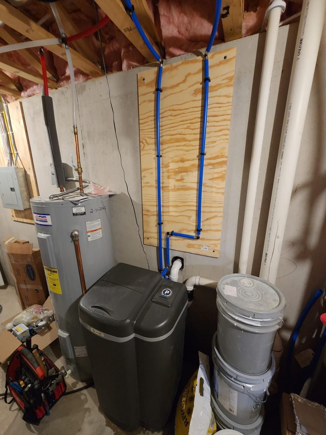 Utility room with water heater, electrical panels, stacked trash bins, and exposed wiring on unfinished walls