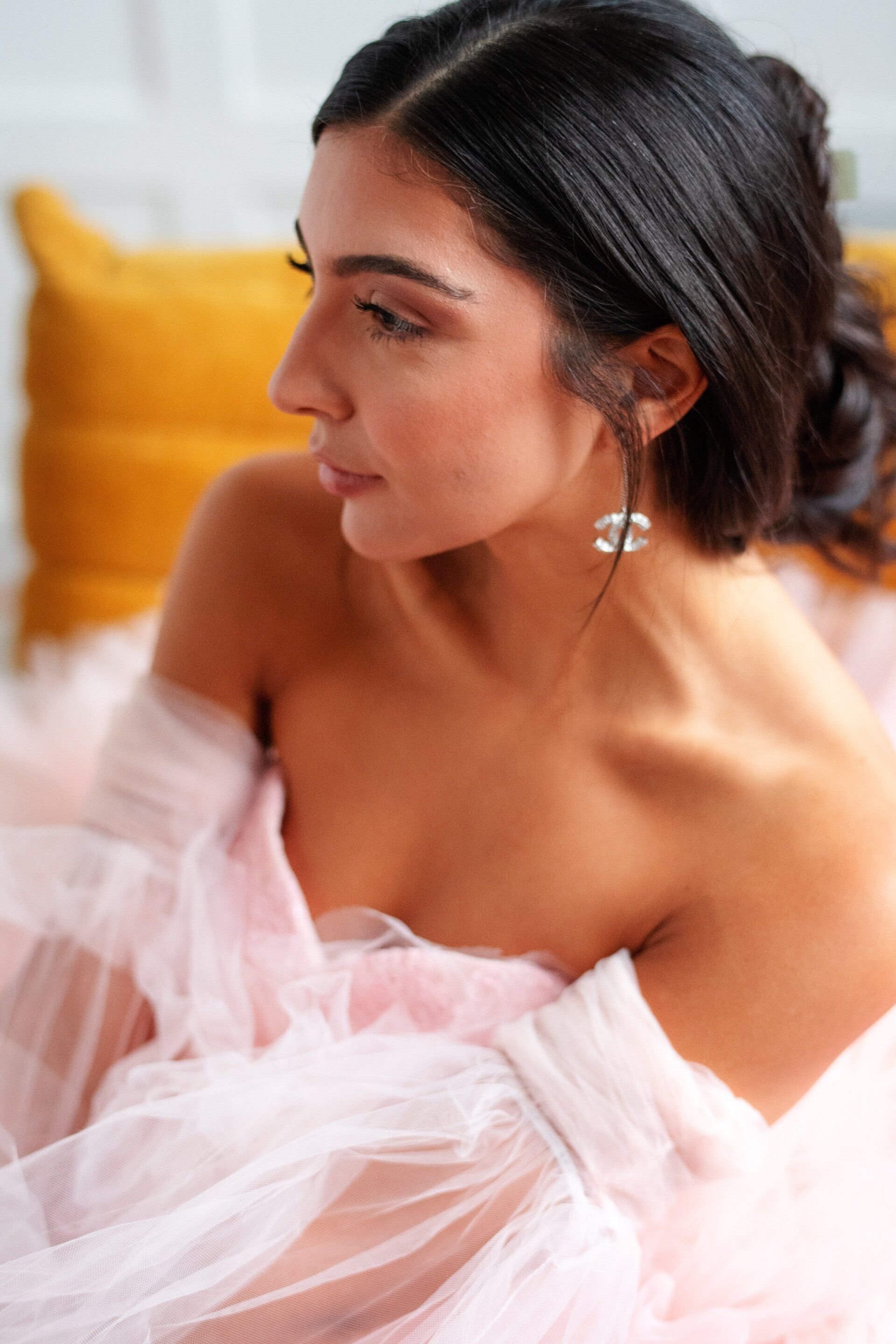 A woman in a pink dress and earrings is sitting on a bed.
