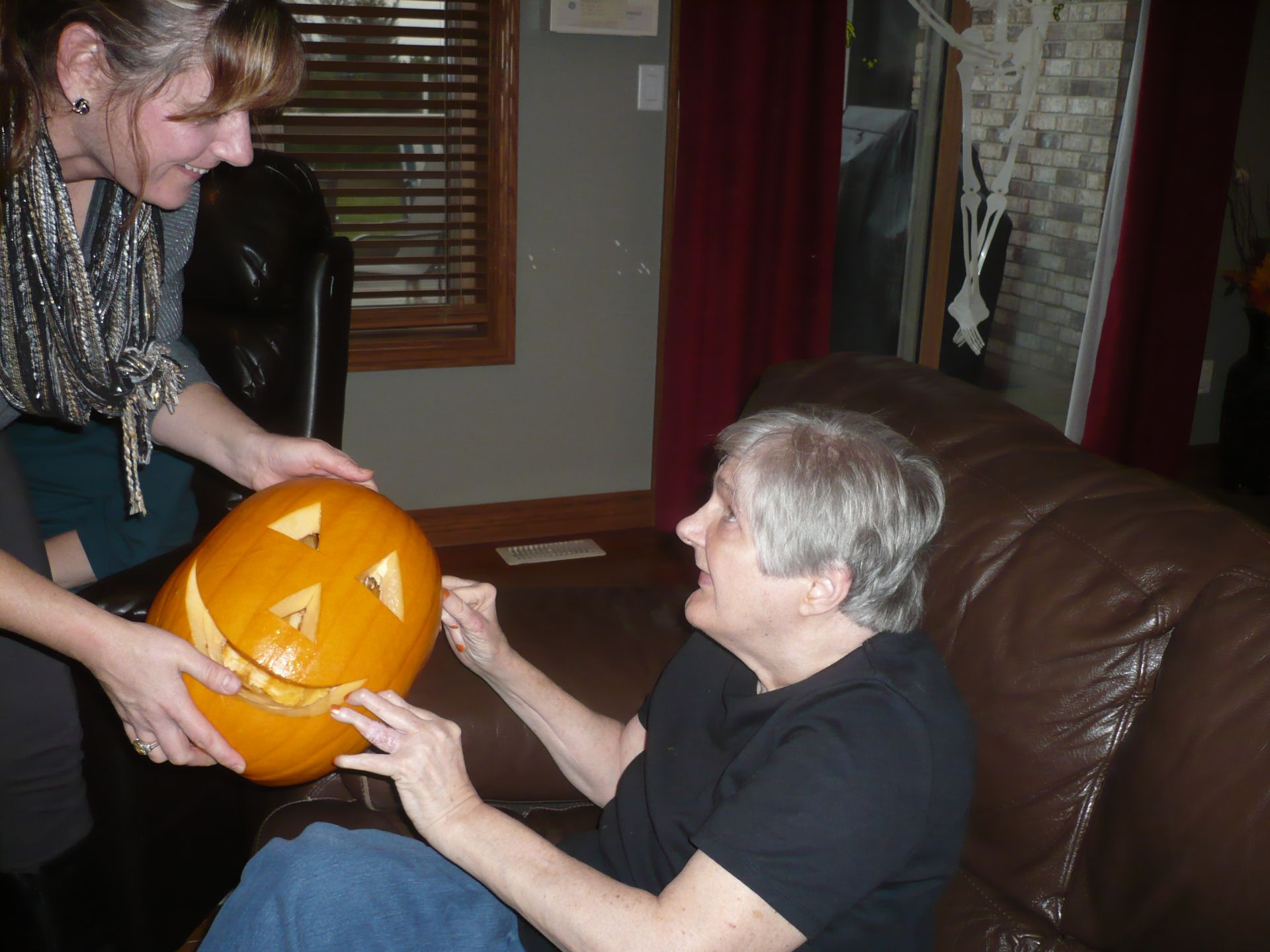 Woman shows a carved pumpkin to another woman on a couch; both smiling.