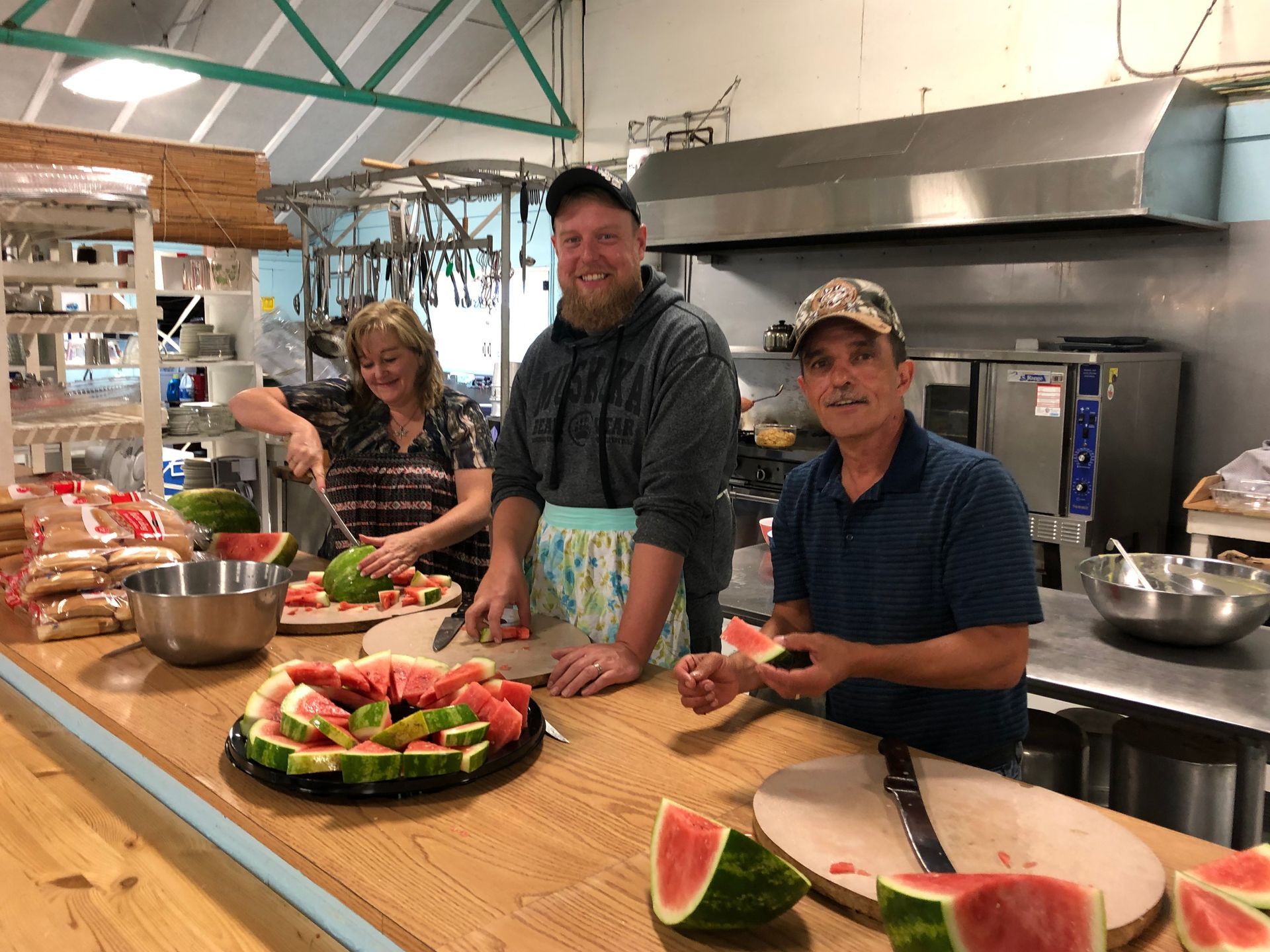 Three people cutting watermelon at a counter in a kitchen.