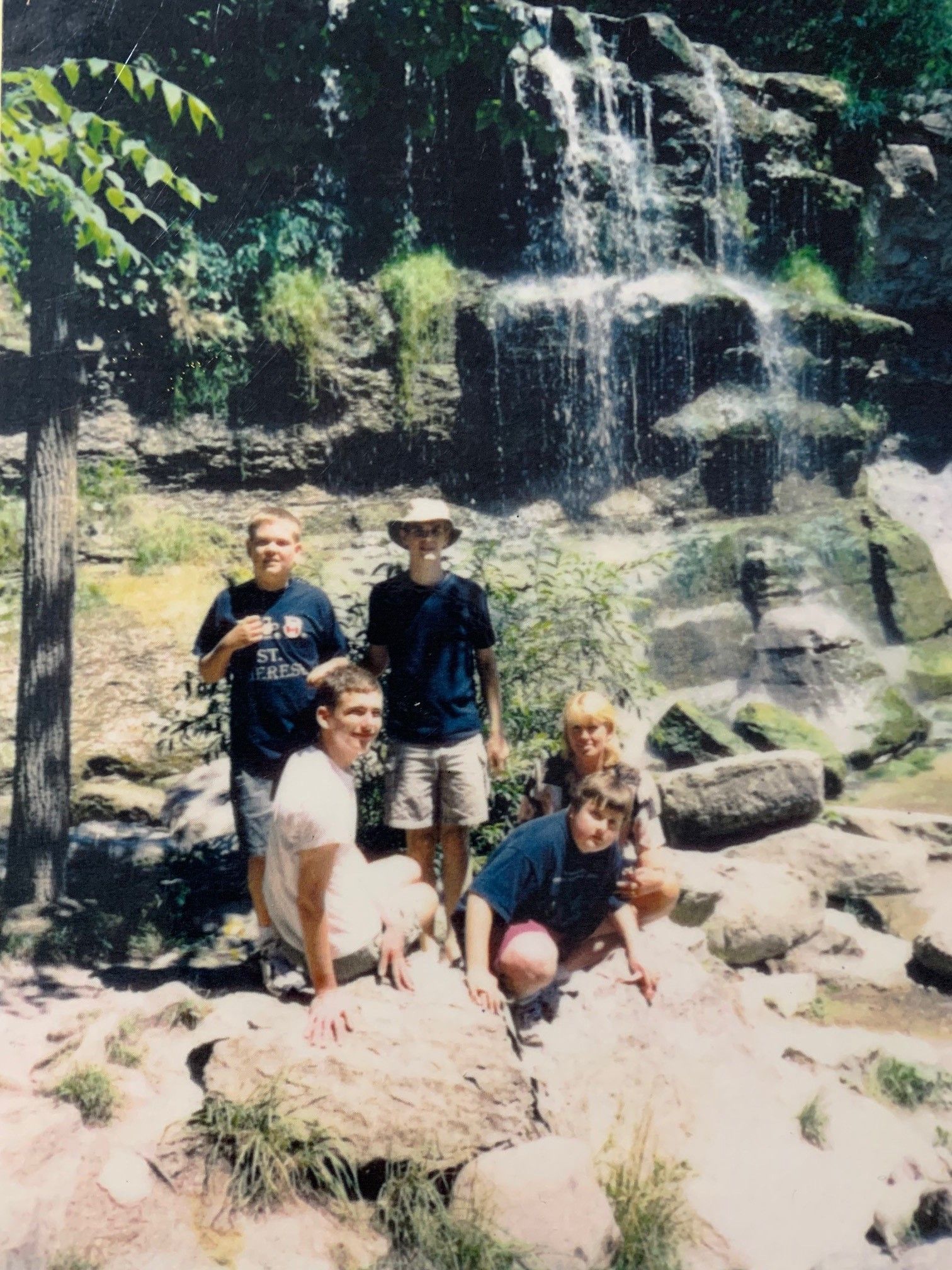 Group of people posing in front of a waterfall. Rocks and trees in the foreground and background.