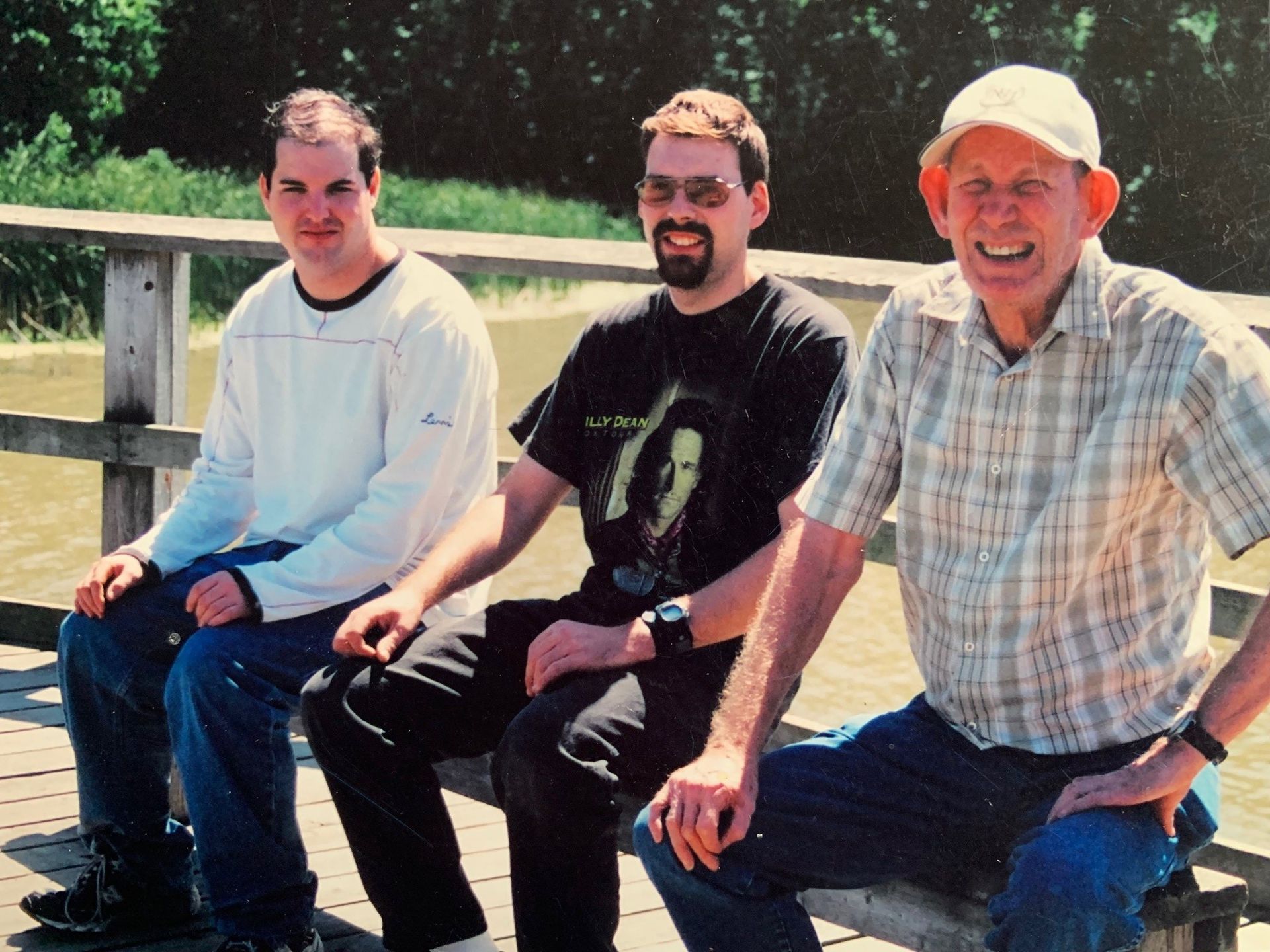 Three men sit on a wooden pier by water. The man on the right smiles; the other two look forward.