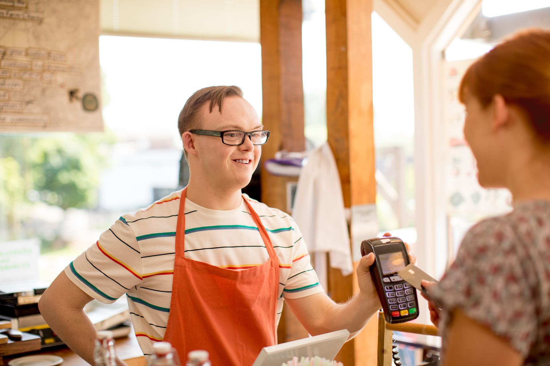 Person in apron smiles while helping a customer pay with a card reader in a shop.