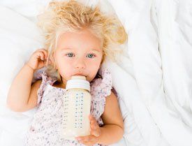 A little girl is drinking milk from a bottle while laying on a bed.